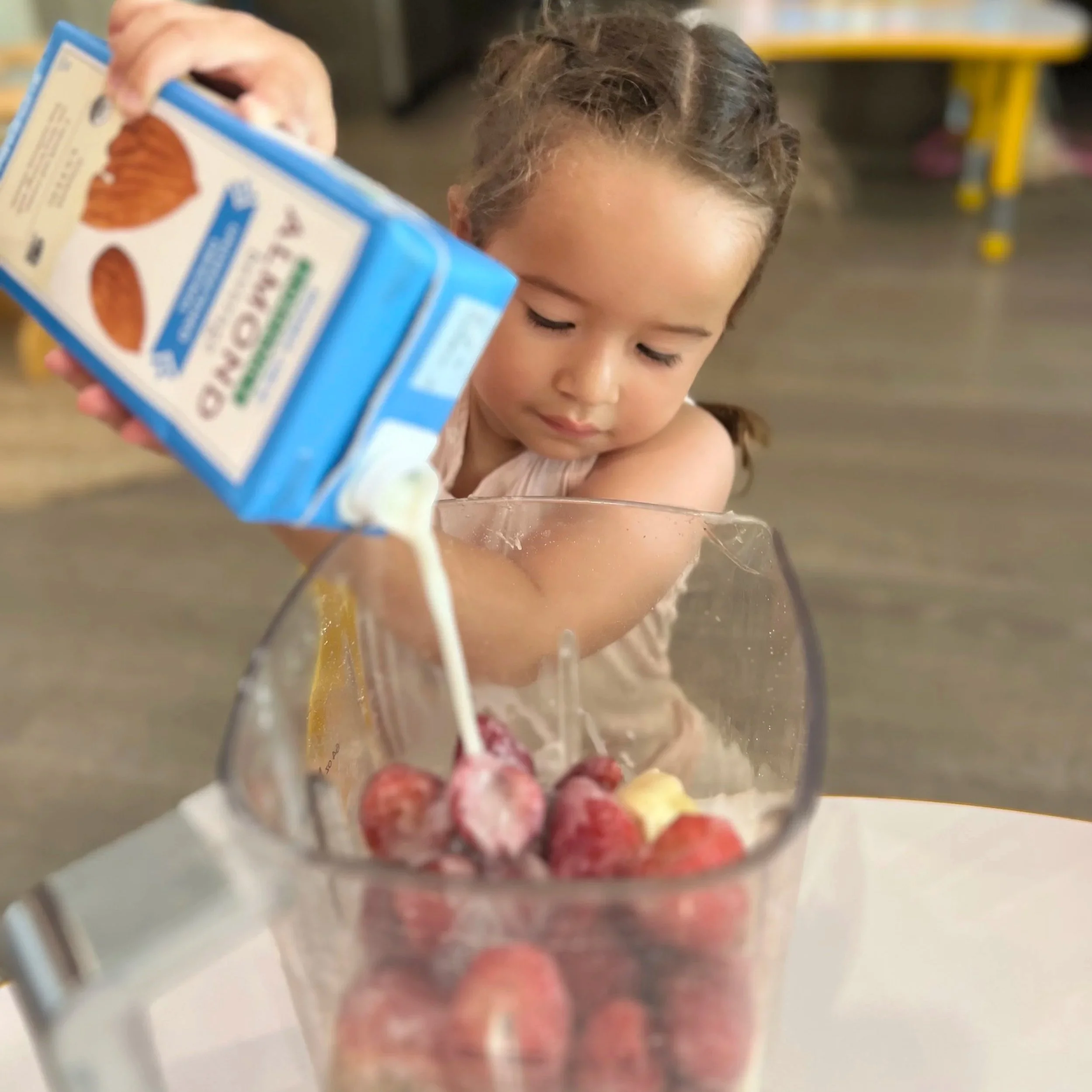 A young girl pouring milk into a blender with strawberries for a smoothie, in a room with yellow and brown furniture.