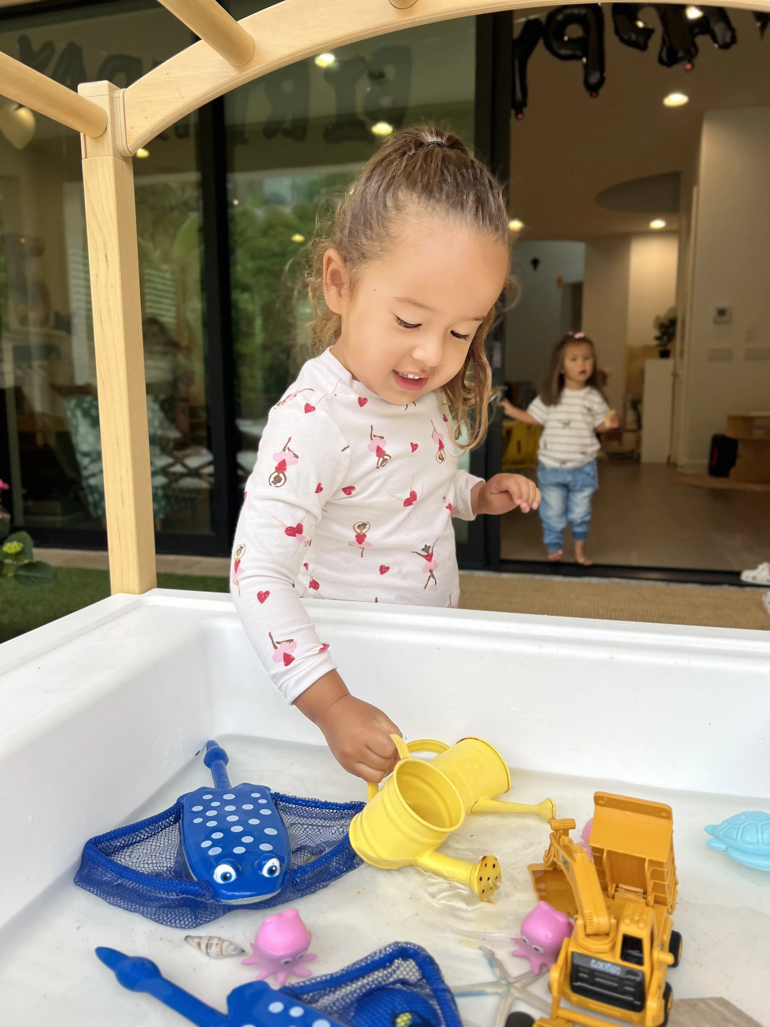 Young girl playing in a sandbox with toy ocean animals and construction vehicles inside a home with another girl in the background.