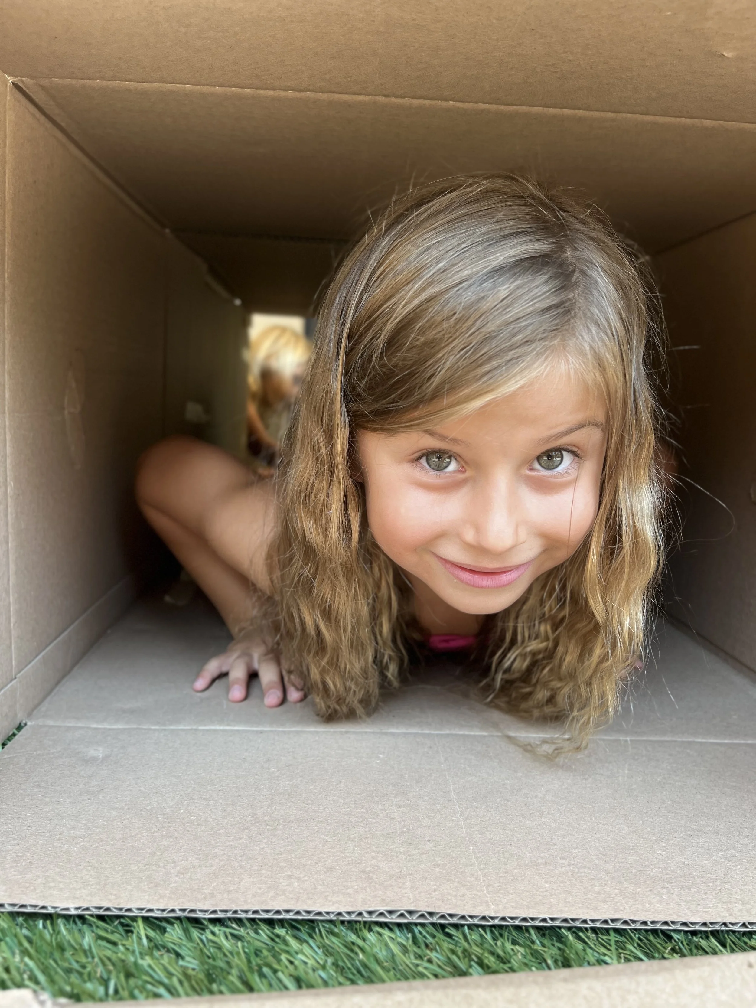 Young girl with long wavy hair crawling through a cardboard box on grass, smiling at the camera.