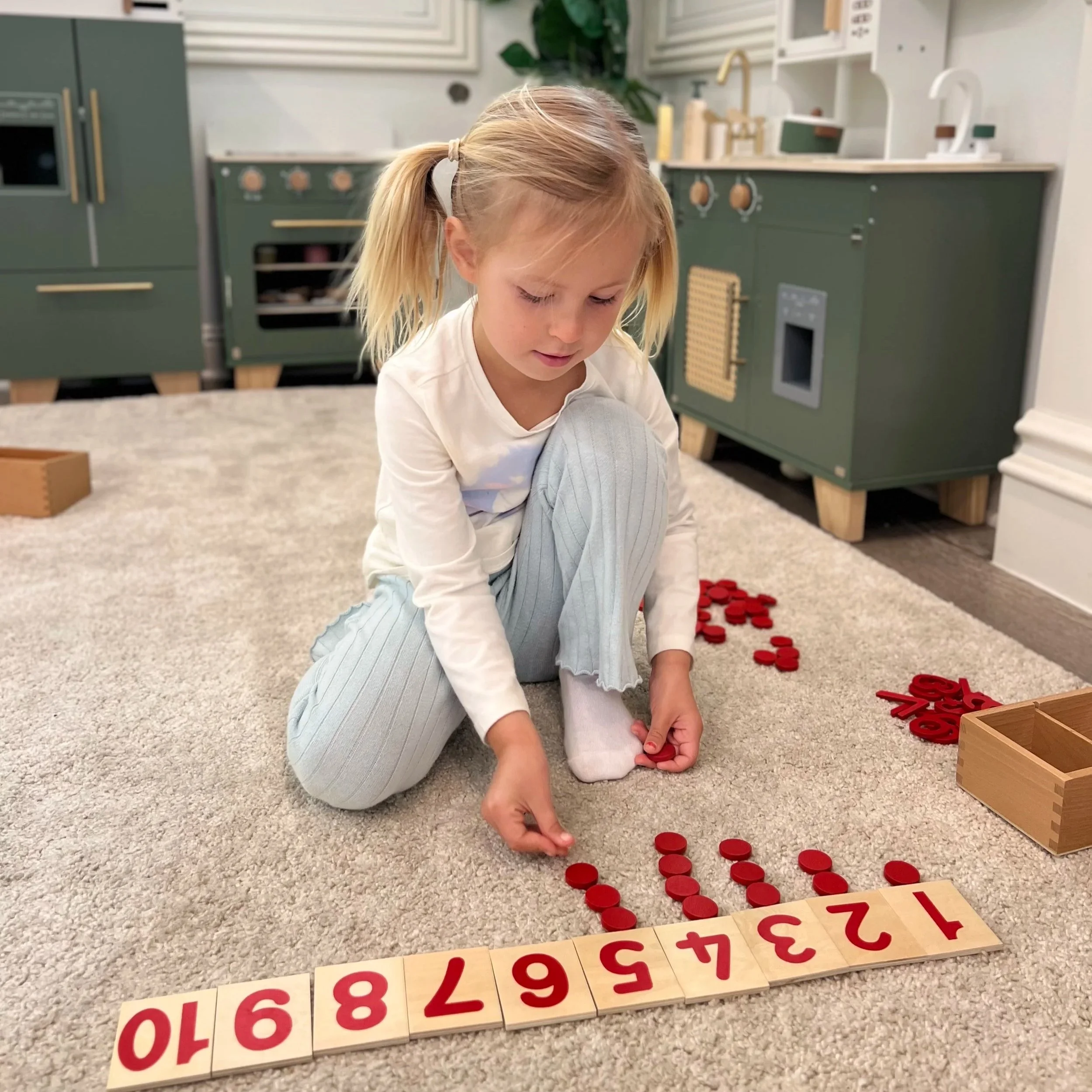 A young girl with blond hair tied in pigtails, kneeling on a carpeted floor, arranging red counters. In front of her, numbered tiles from 1 to 10 are laid out. The background shows a kitchen with green cabinets and various kitchen appliances.