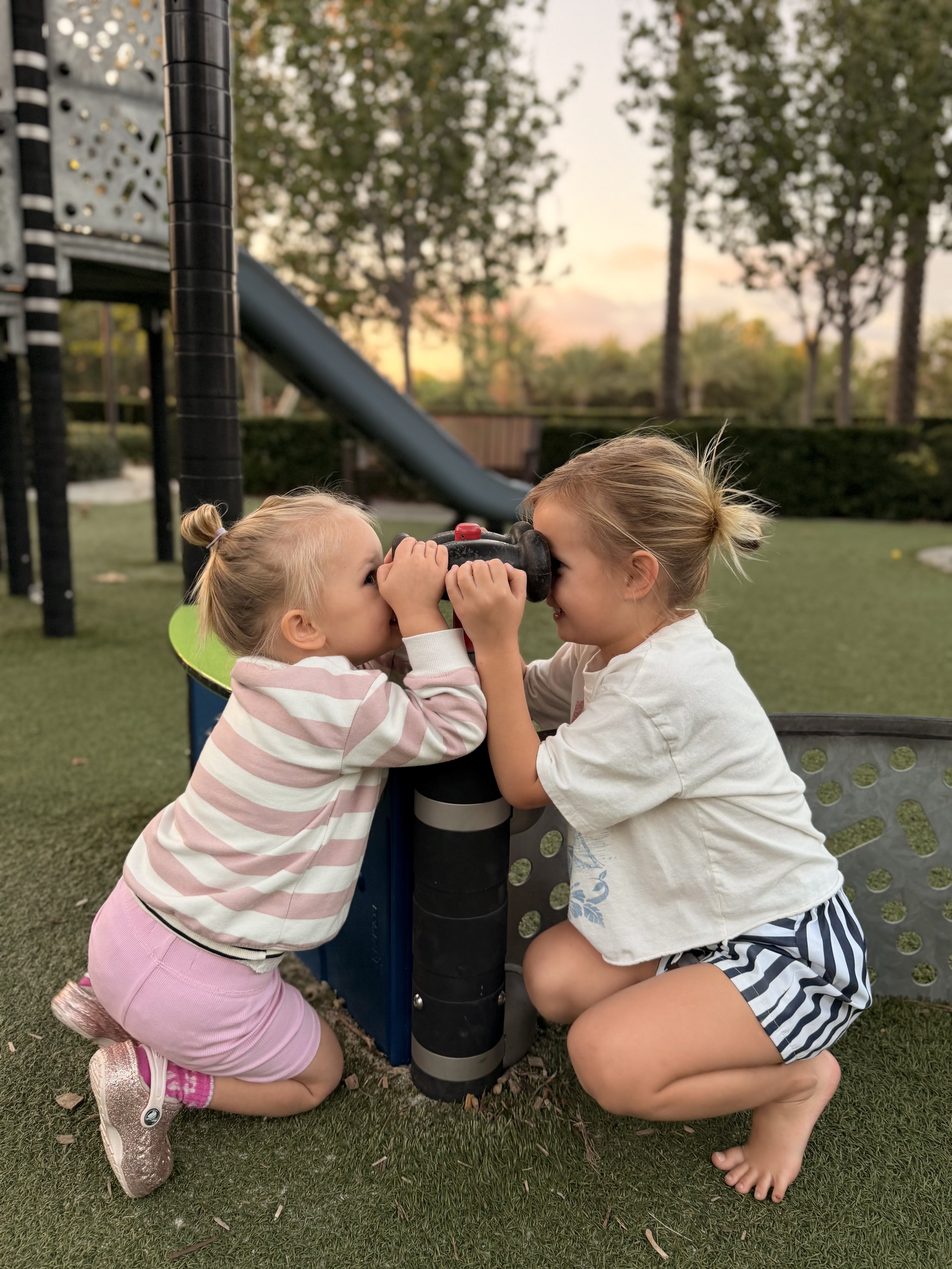 Two young girls, one wearing a pink and white striped hoodie with pink shorts and the other in a white t-shirt with blue and white striped shorts, are playing with a toy telescope outdoors during sunset on a grassy playground.