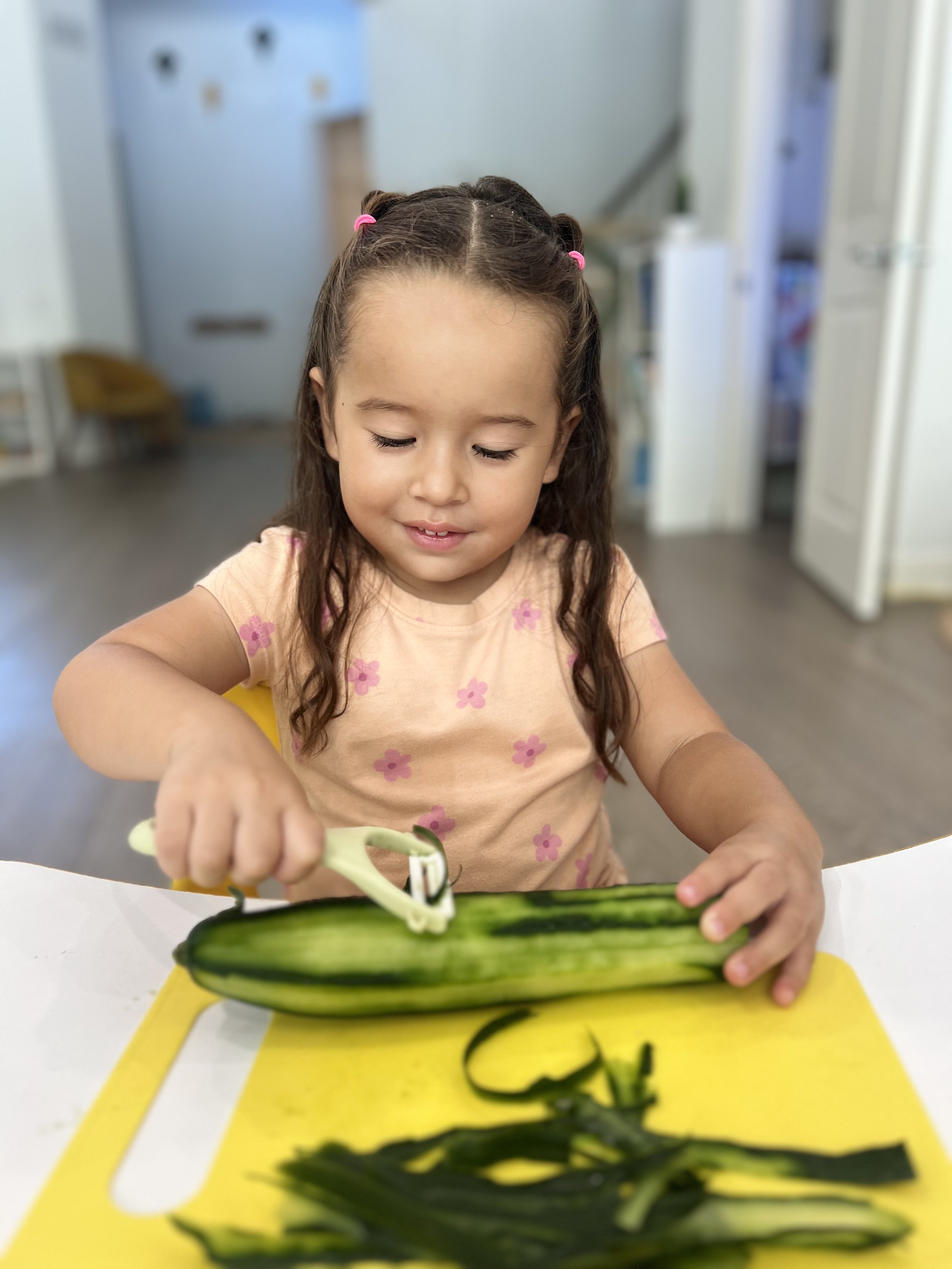 Young girl peeling a cucumber with a vegetable peeler on a white table with a yellow cutting board.