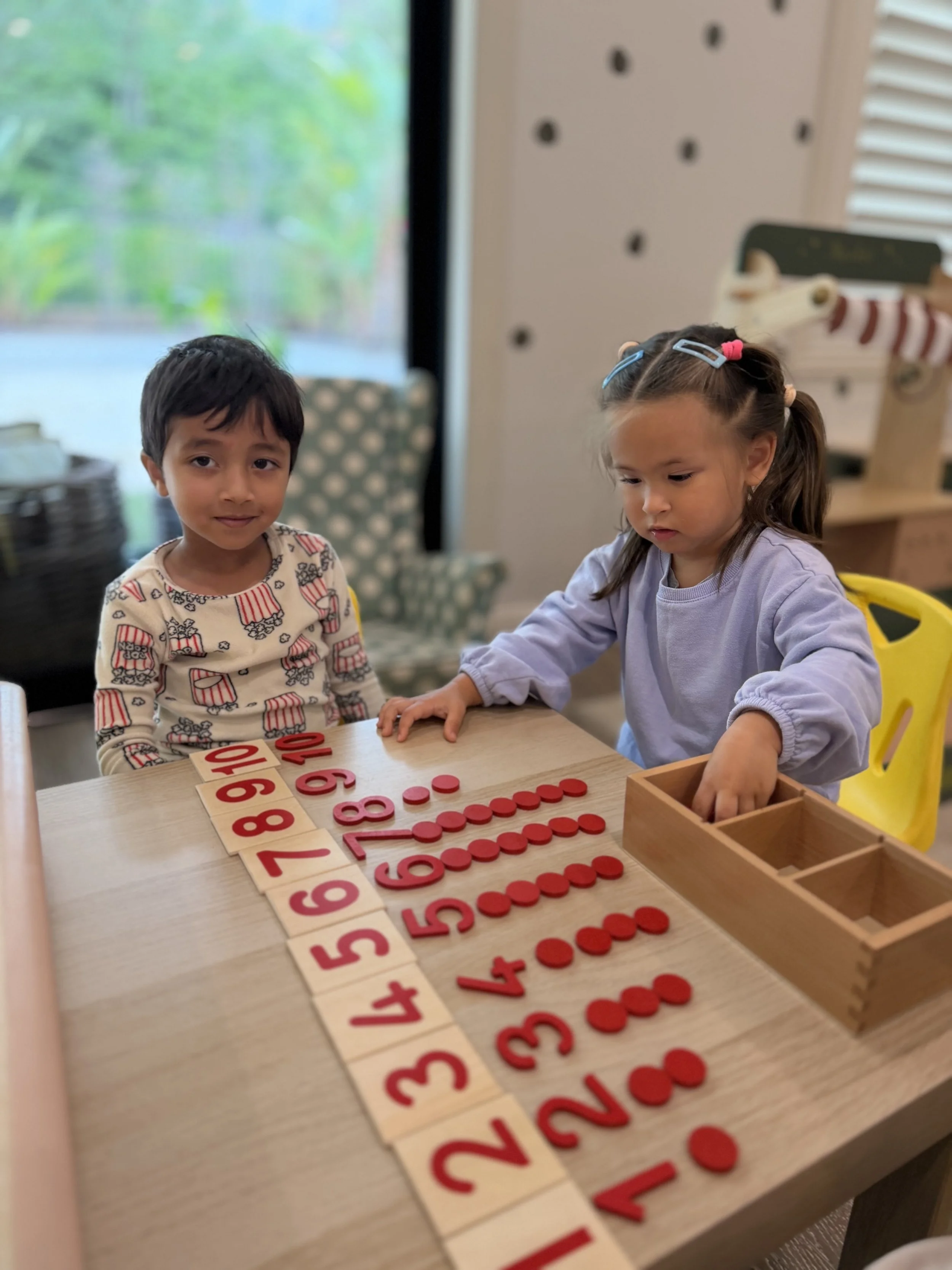 Two young girls playing with number tiles at a table indoors, with one girl looking at the camera and the other reaching into a wooden box.