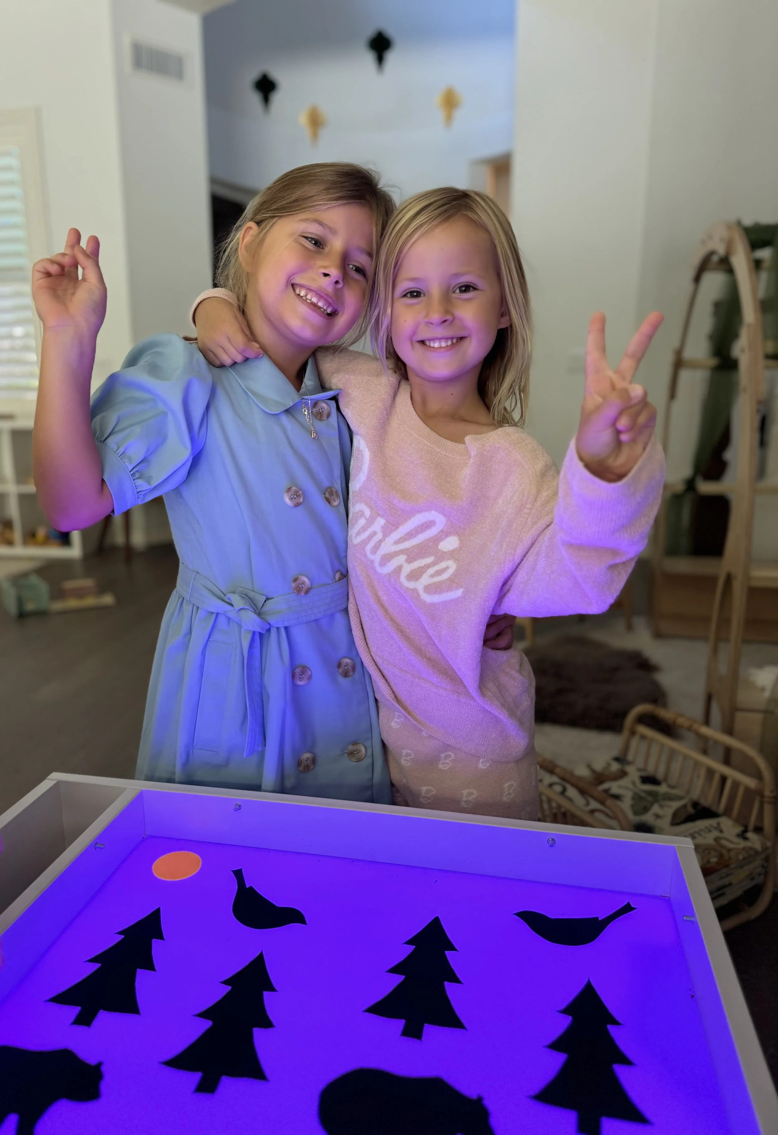 Two young girls are smiling and posing for a photo, with one girl wearing a light blue dress and the other a light pink shirt that says 'breathe.' They are in a cozy room with a dark wooden floor, a white wall with black and gold wall decorations, and a glowing purple lightbox with Halloween-themed cutouts of trees, bats, and a pumpkin.