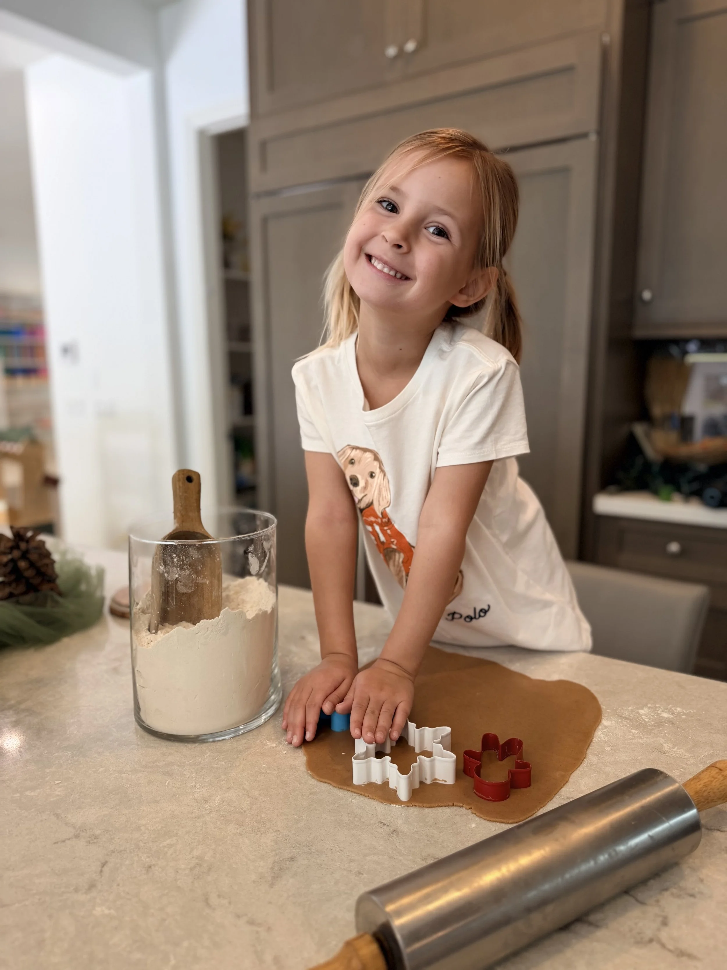 A smiling young girl with light skin and red hair in pigtails, wearing a white t-shirt with a dog graphic, is baking cookies in the kitchen. She is pressing cookie cutters into rolled-out gingerbread dough on a kitchen counter, with a flour sifter and rolling pin nearby.
