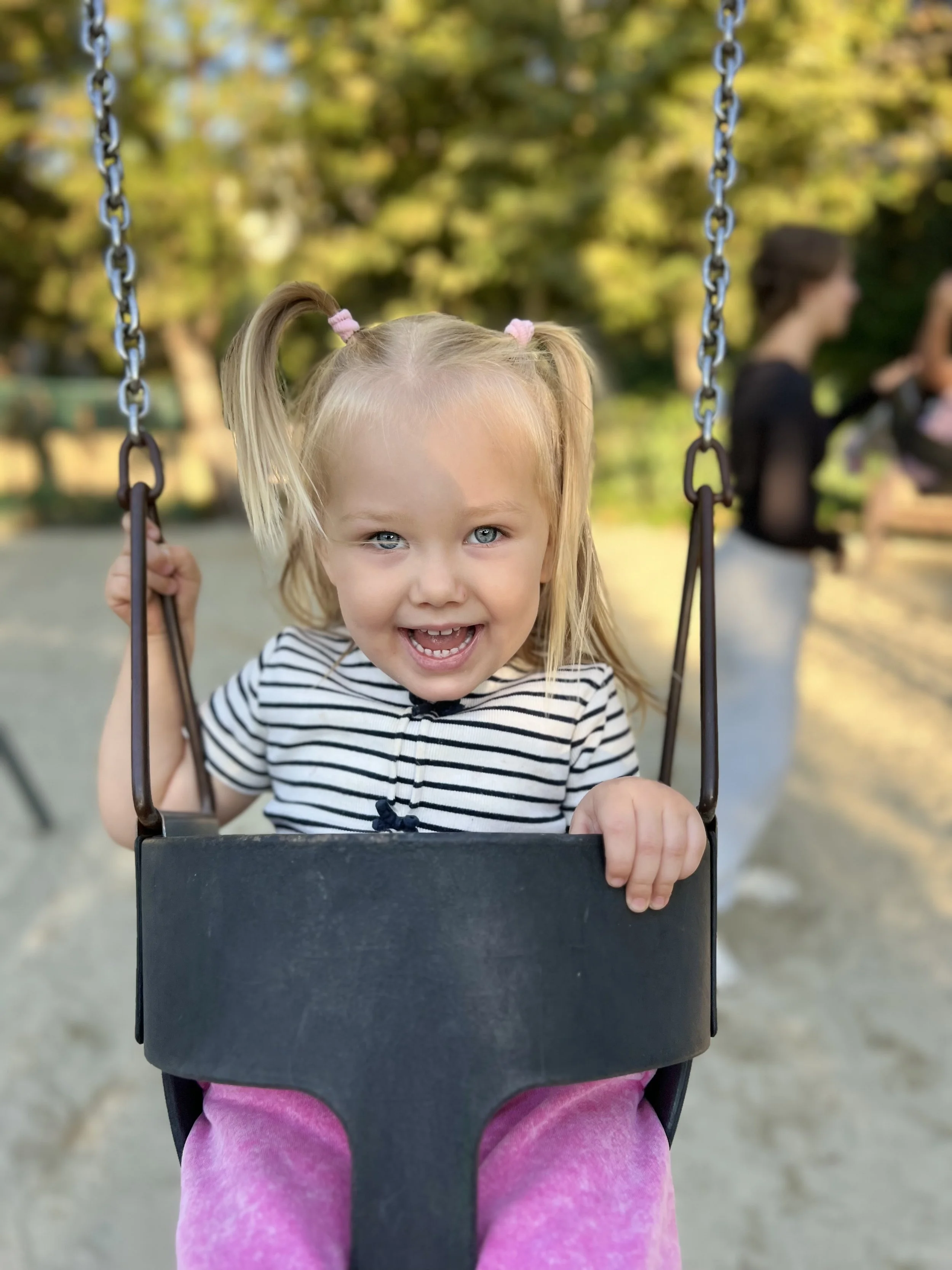 A young girl with blonde hair in pigtails, wearing a striped shirt and pink pants, smiling while sitting in a playground swing.