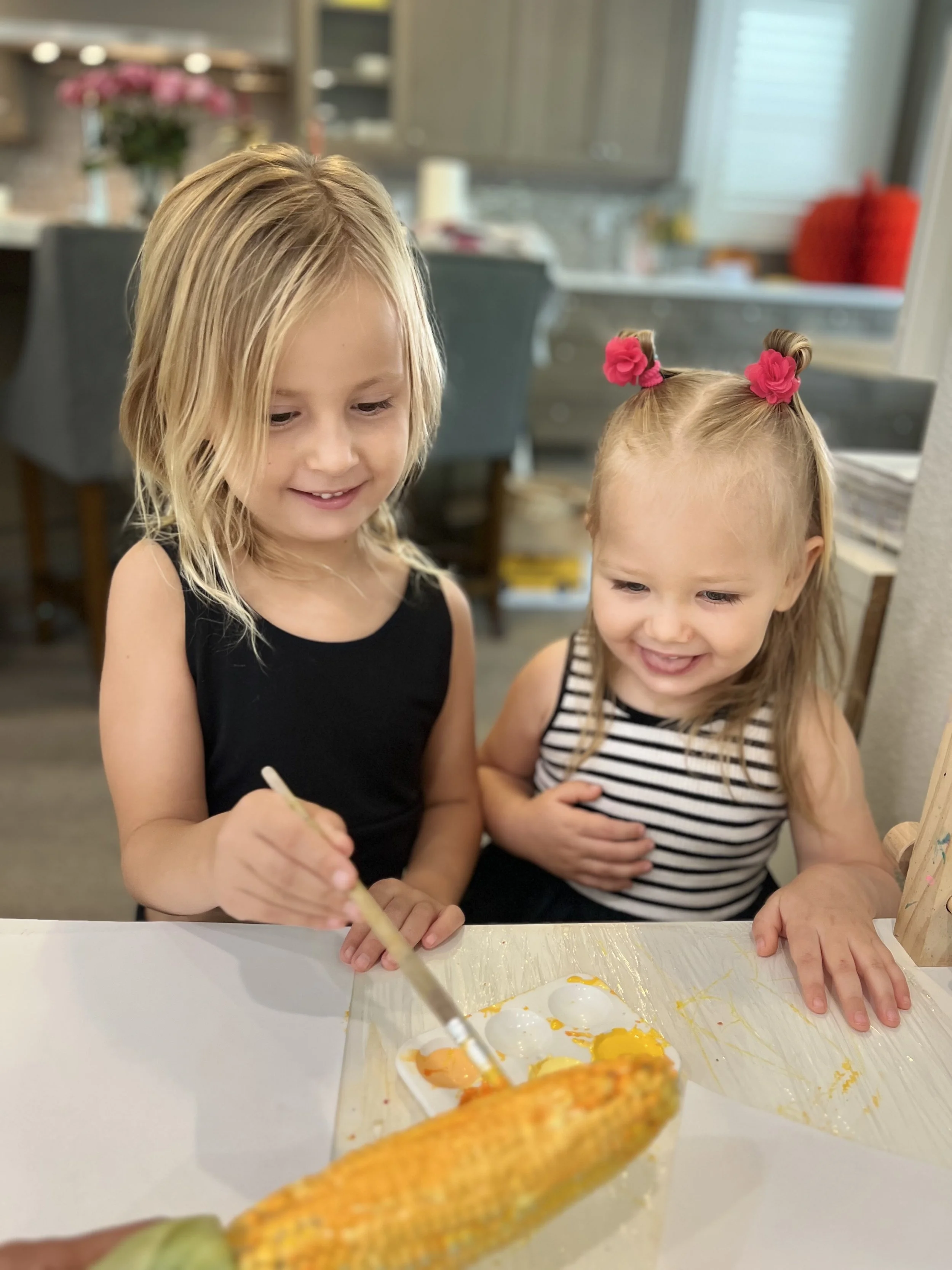 Two young girls smiling and watching as one shucks corn over a tray with eggs in a kitchen.