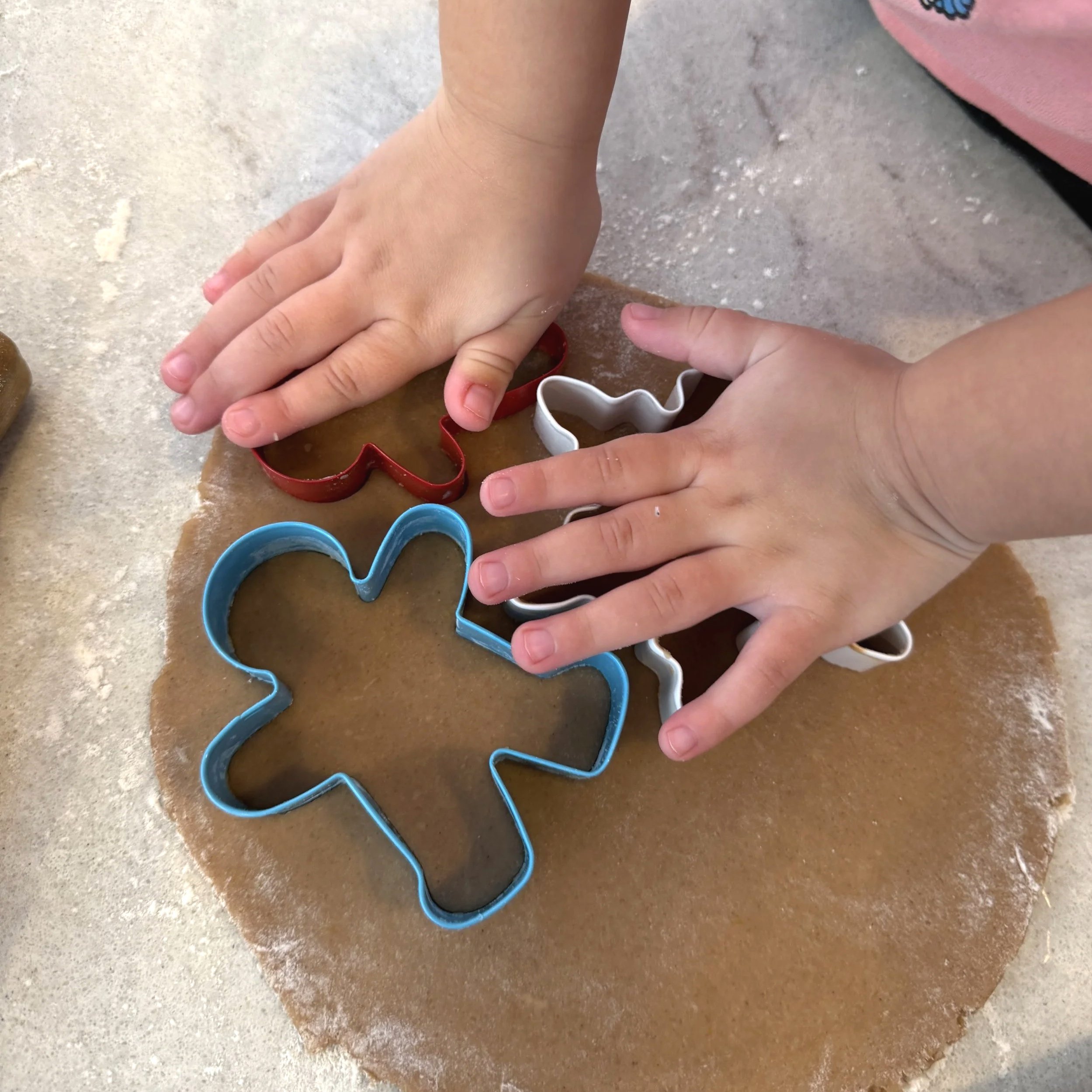 Two children’s hands pressing cookie cutters into rolled brown dough on a floured surface.