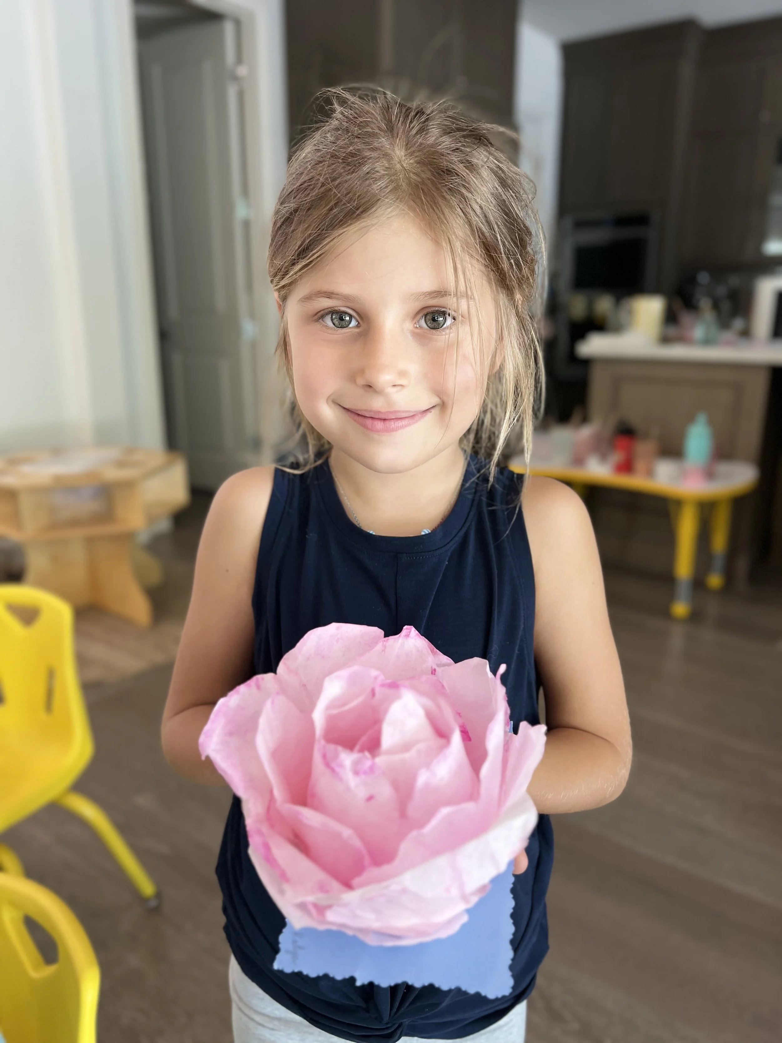 A young girl with light brown hair and blue eyes smiling while holding a pink paper flower in a blue holder, standing inside a room with yellow chairs and a kitchen in the background.