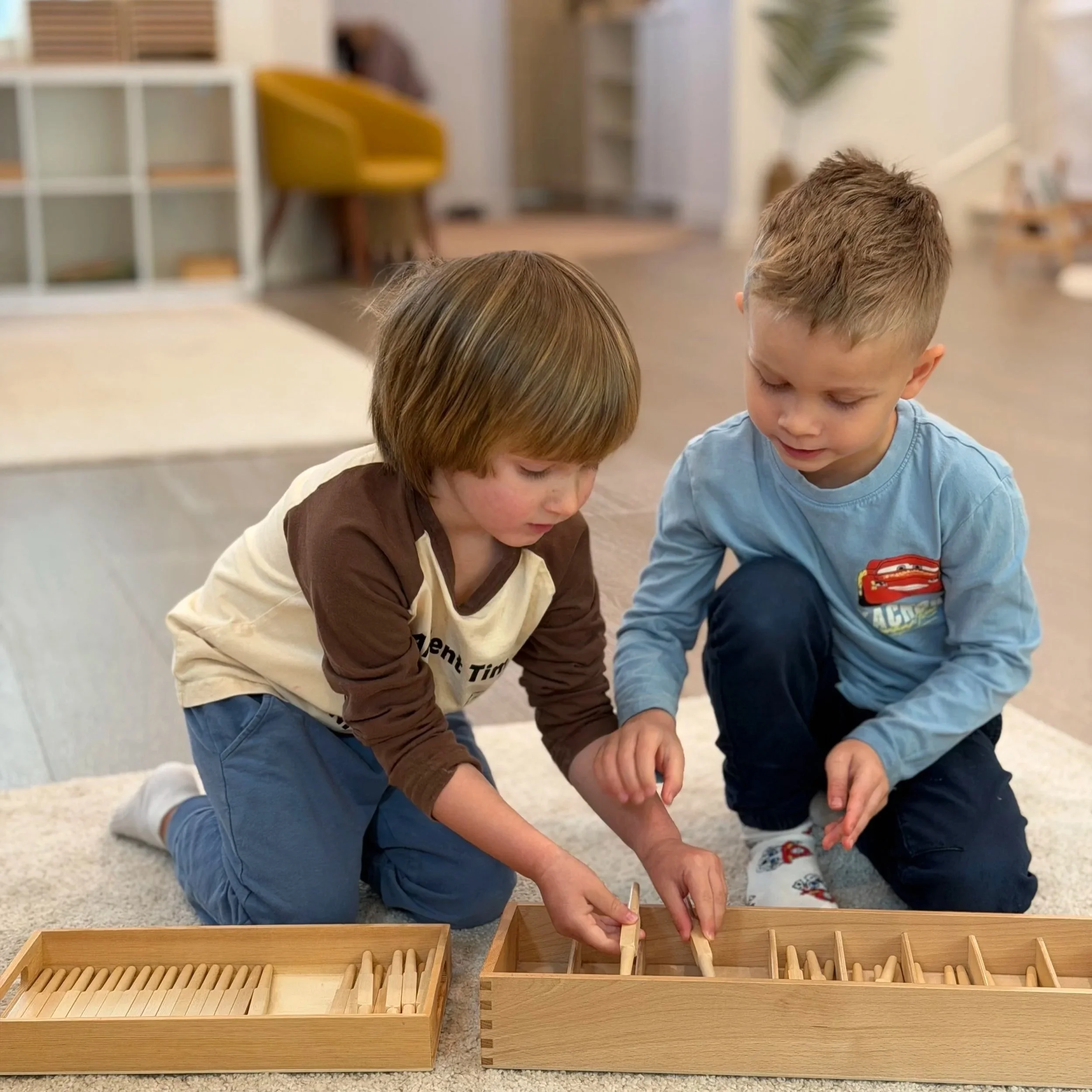 Two young boys are sitting on a carpeted floor, playing with wooden dominoes stored in wooden trays.
