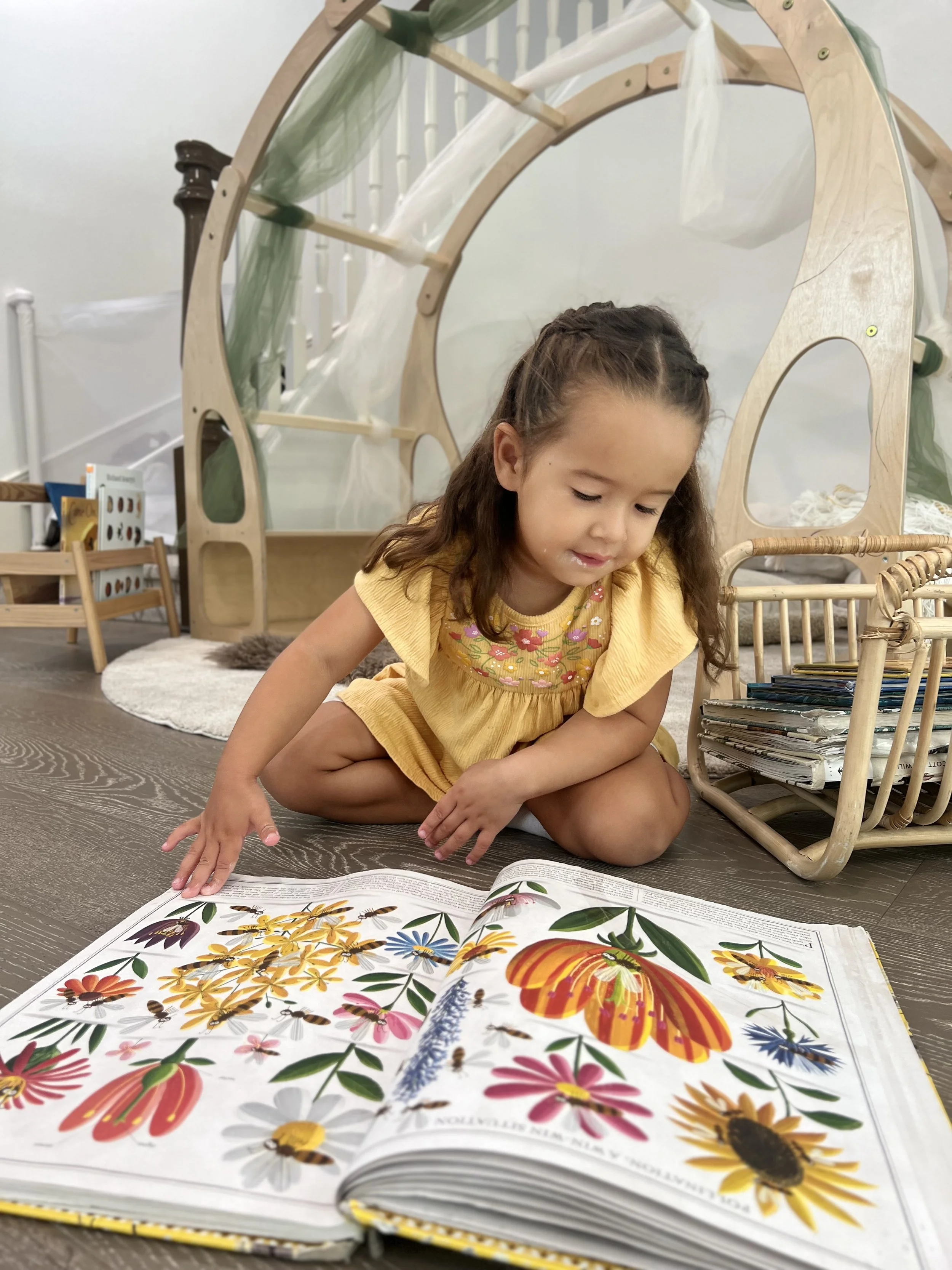 Young girl with curly dark hair in a yellow dress sitting on a wooden floor, looking at a large colorful book about flowers and insects with bees on it, in a cozy indoor setting with a wooden playhouse in the background.