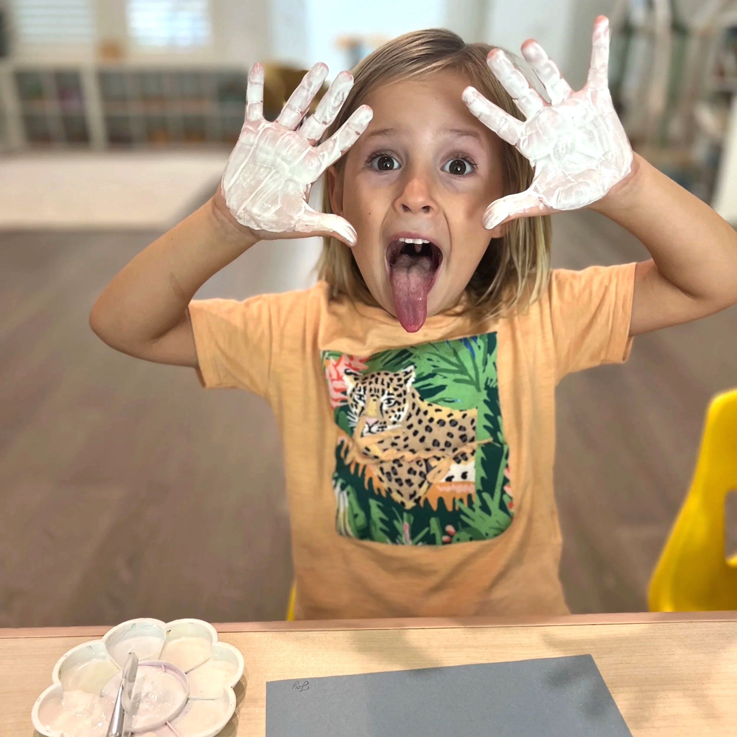 A young girl with blonde hair making a silly face with her tongue out and eyes wide open, holding her hands with white paint on them up near her face, sitting at a table with paint palettes and paper.