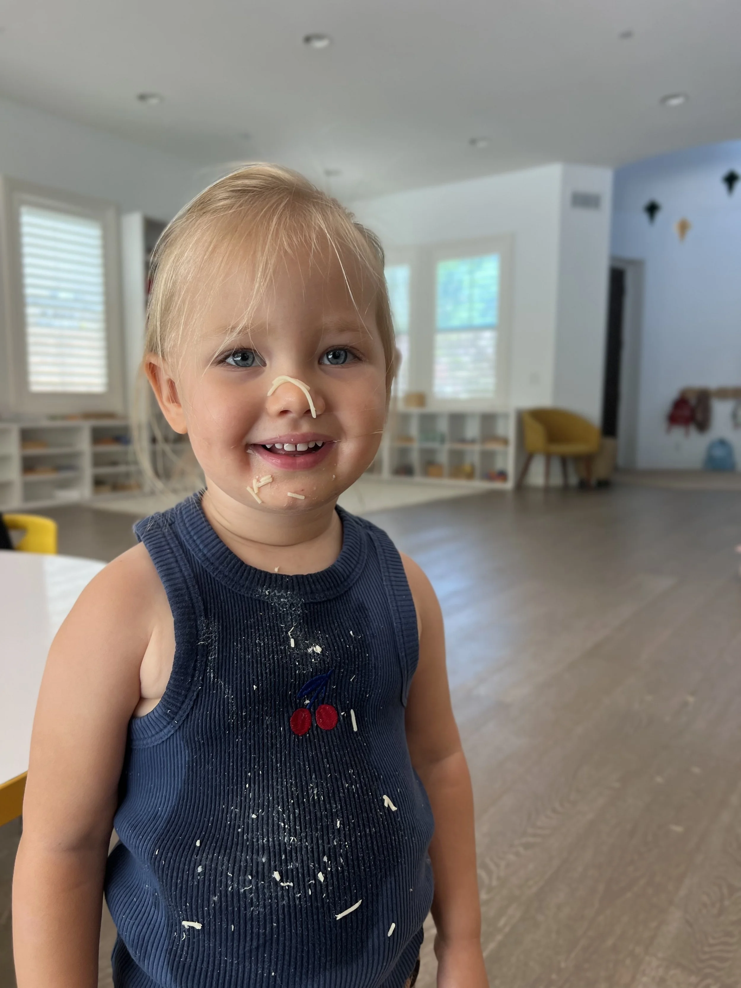 A young child with blonde hair and blue eyes, wearing a dark tank top with a cherry design, smiling with food on face and shirt inside a bright, playroom with windows and shelves.
