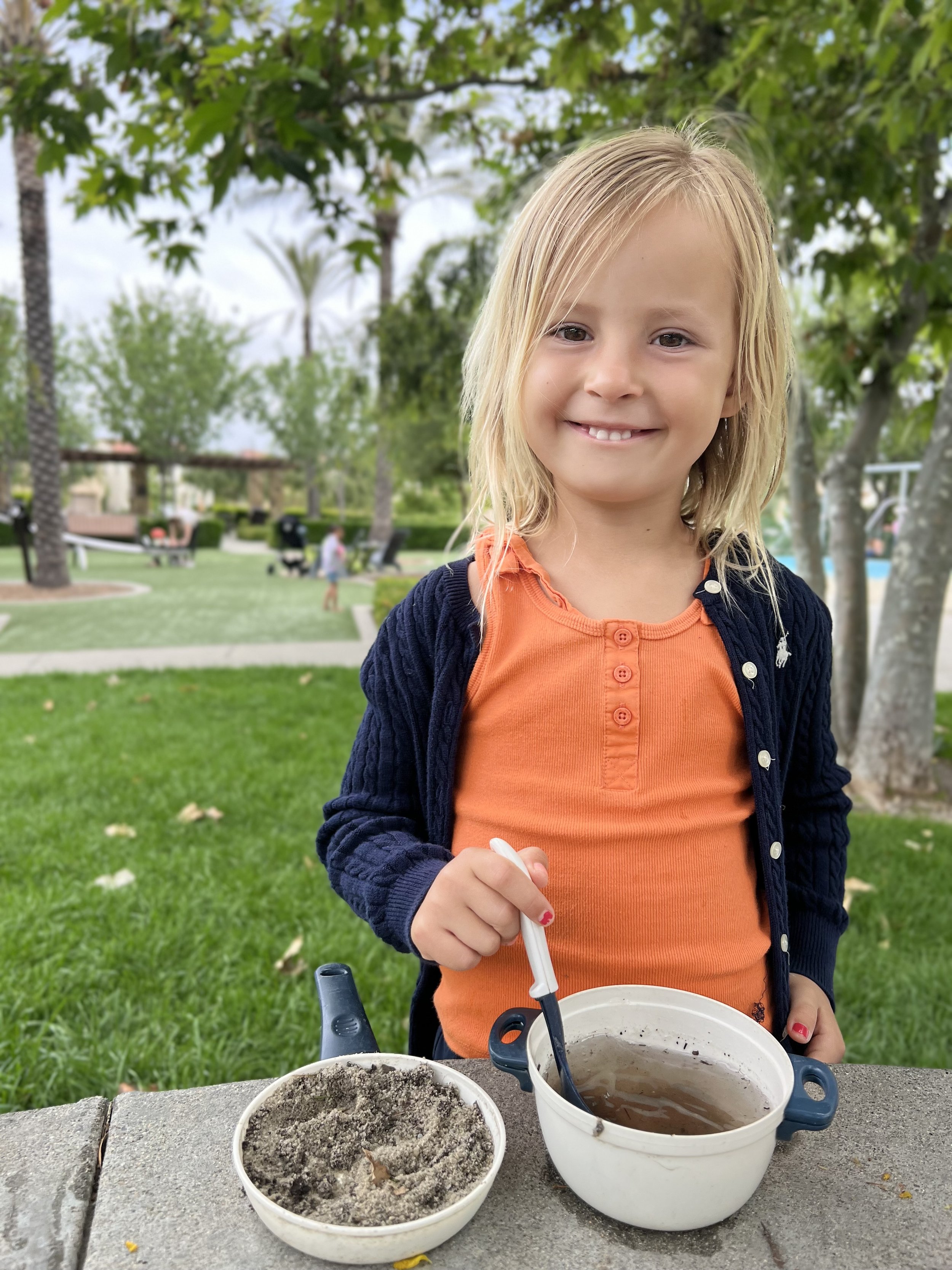A young girl with long blonde hair, wearing an orange top and a navy blue sweater, stands outdoors at a park, smiling at the camera. She is holding a spoon and has two bowls in front of her on a stone table; one bowl contains dirt and the other has a liquid, possibly mud or water.