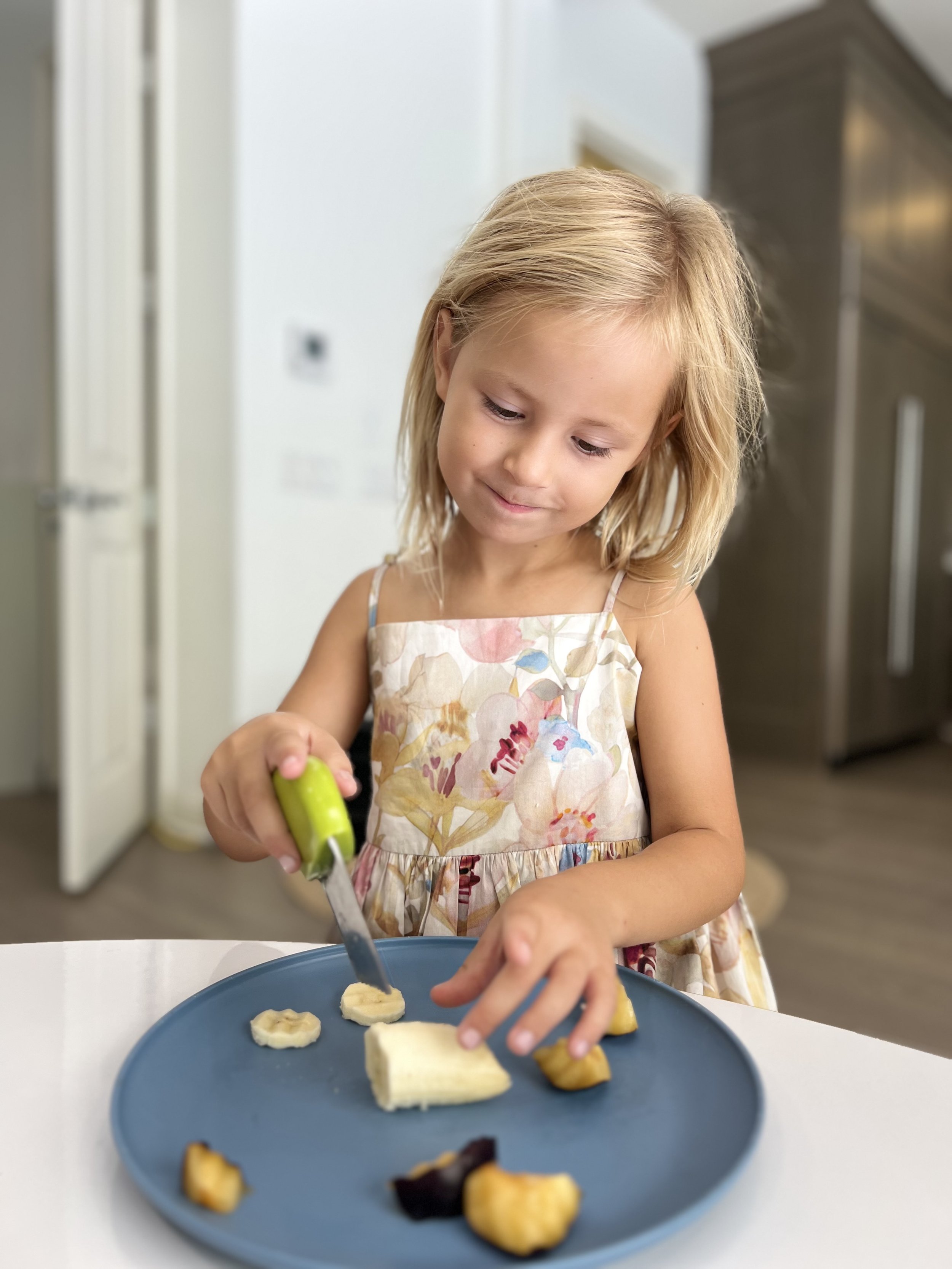 A young girl with blonde hair wearing a floral dress is cutting banana slices on a blue plate with a small green-handled knife in a kitchen.