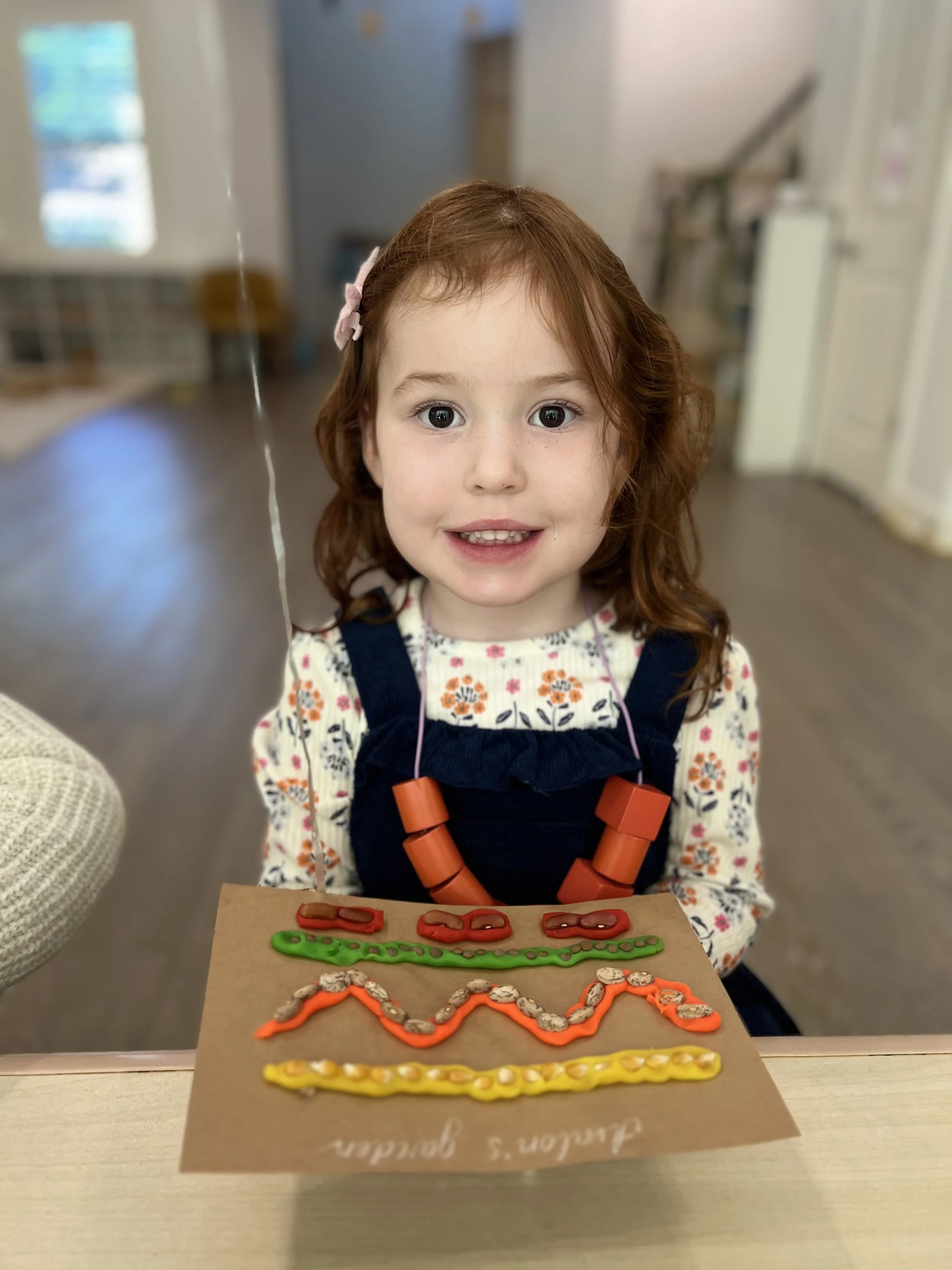 A young girl with red hair and a floral shirt, wearing a navy dress and orange necklace, smiling and showing decorated pretzels and snacks on a brown paper.