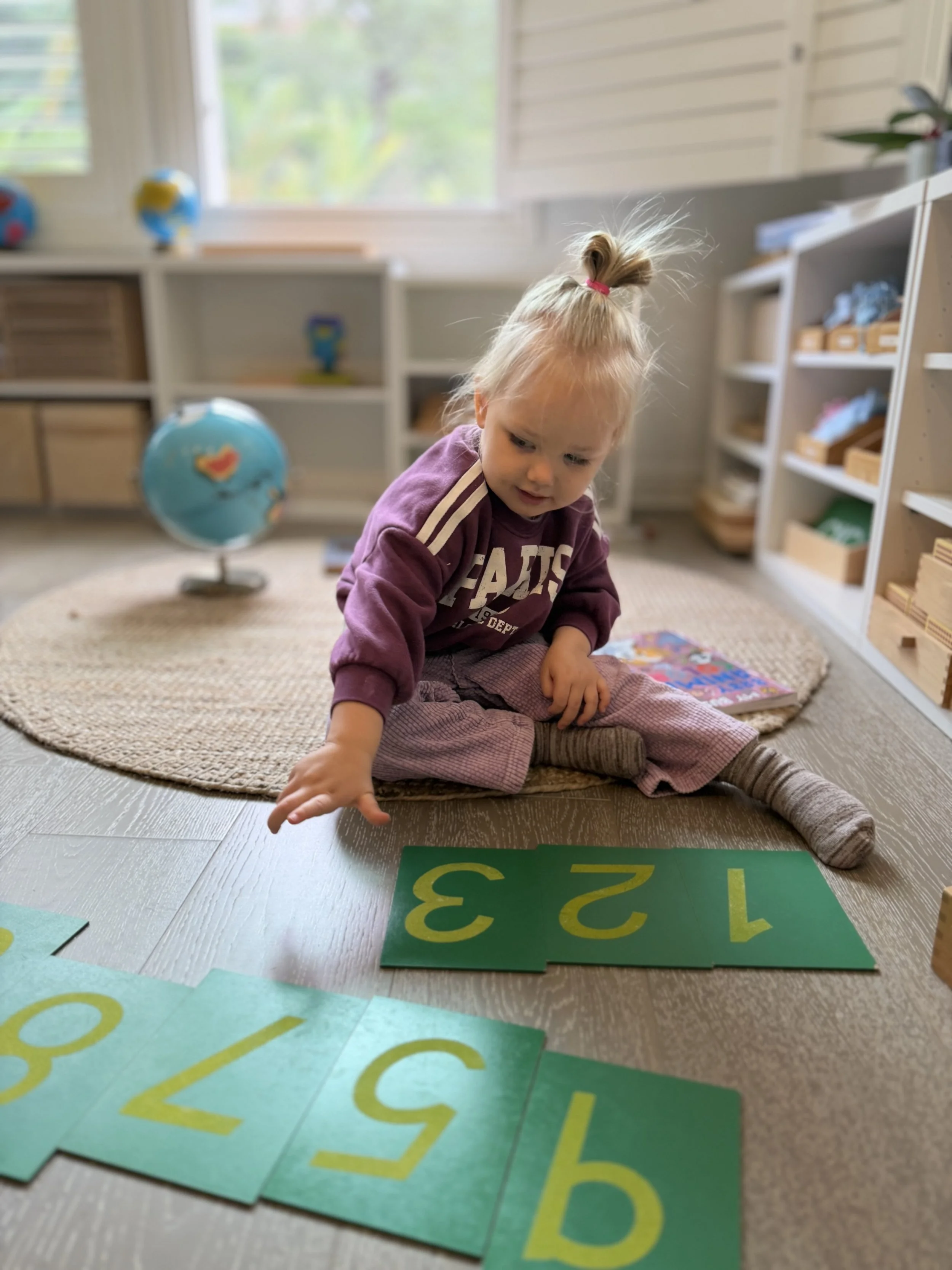 A young girl with blonde hair tied up in a ponytail, sitting on the floor in a classroom or playroom, sorting large green flashcards with yellow numbers and letters on them.