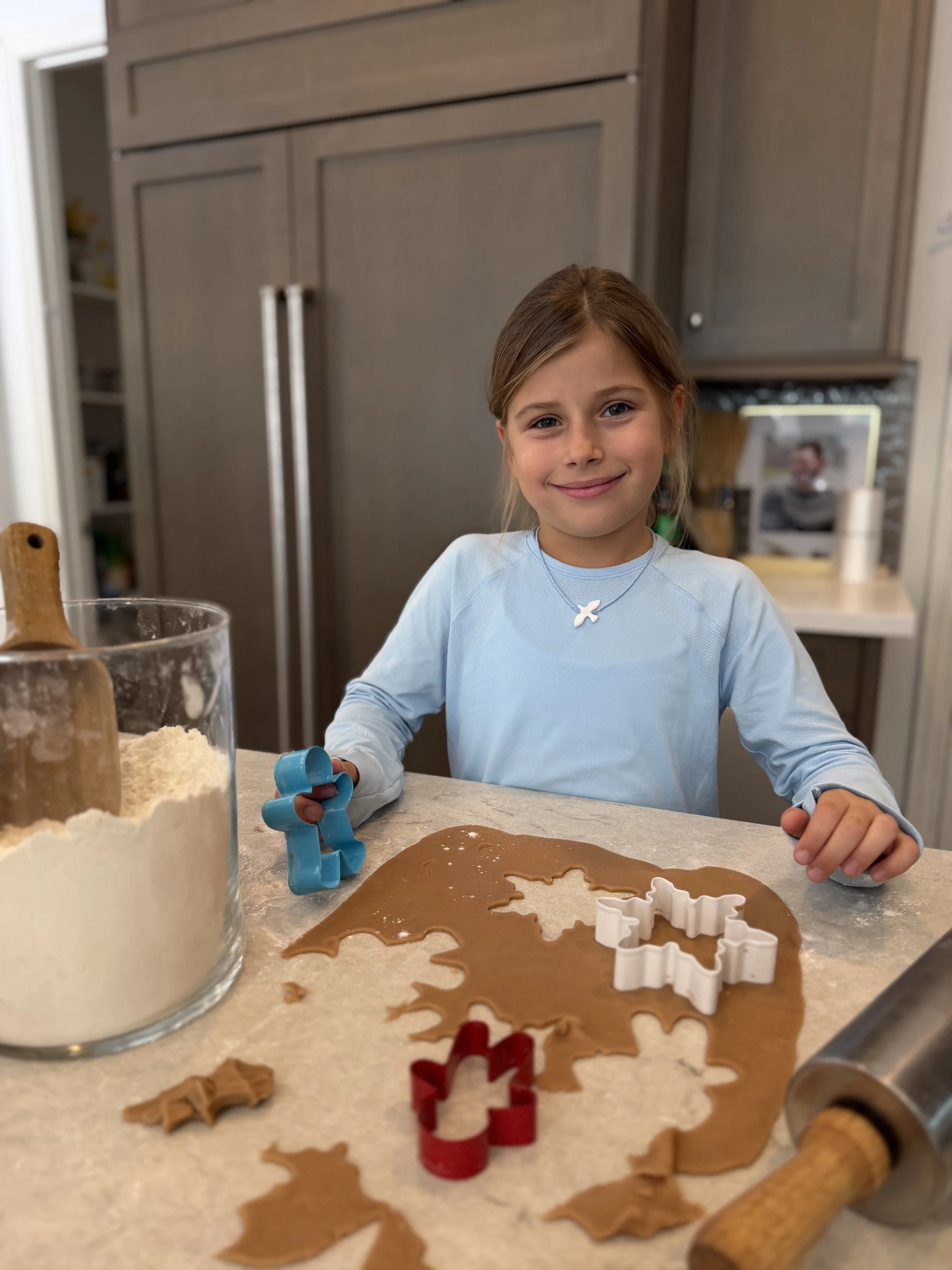 A young girl in a blue long-sleeve shirt smiling at the camera while making holiday cookies with cookie cutters on gingerbread dough in a kitchen.