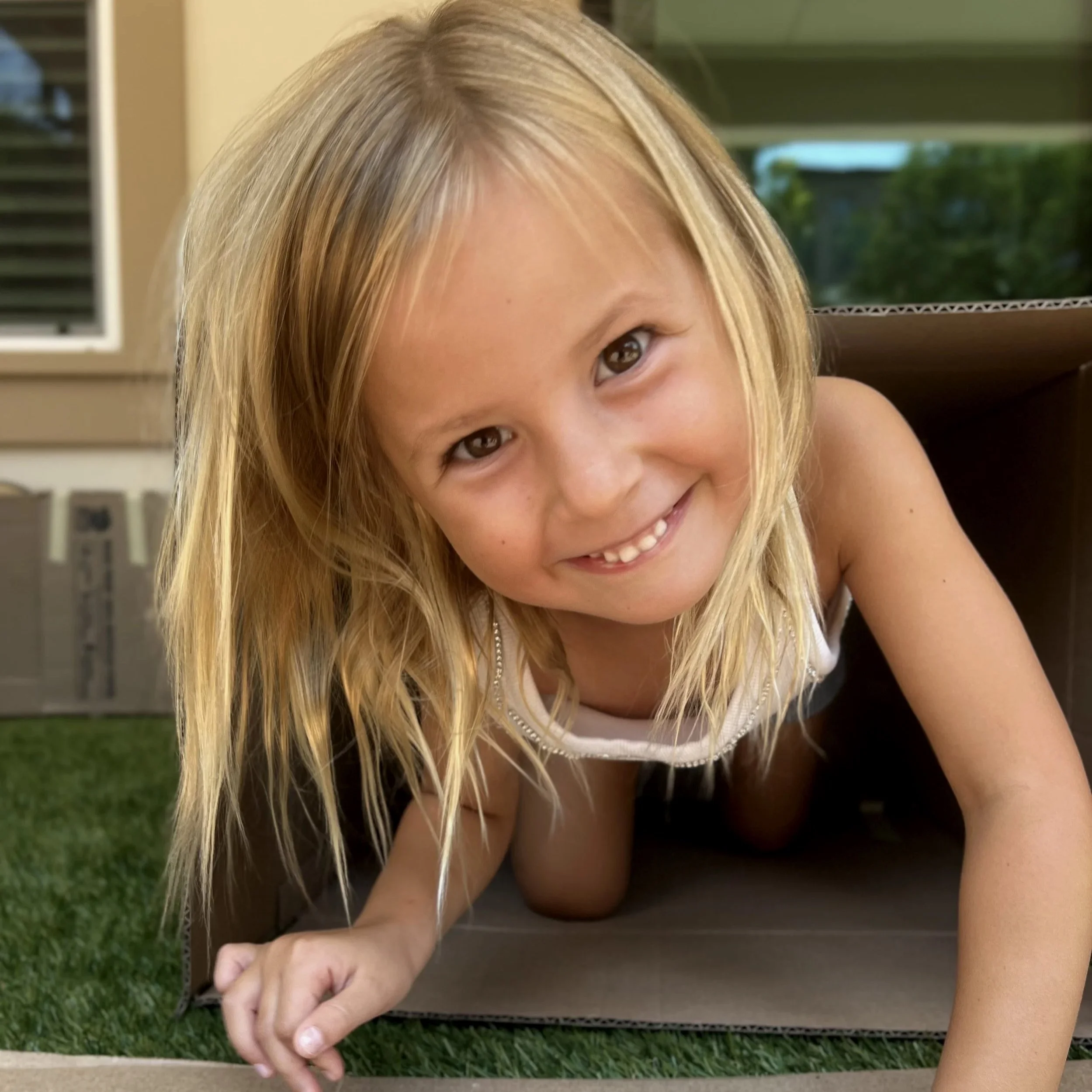 A young girl with long blonde hair smiling while crawling out of a cardboard box outdoors.