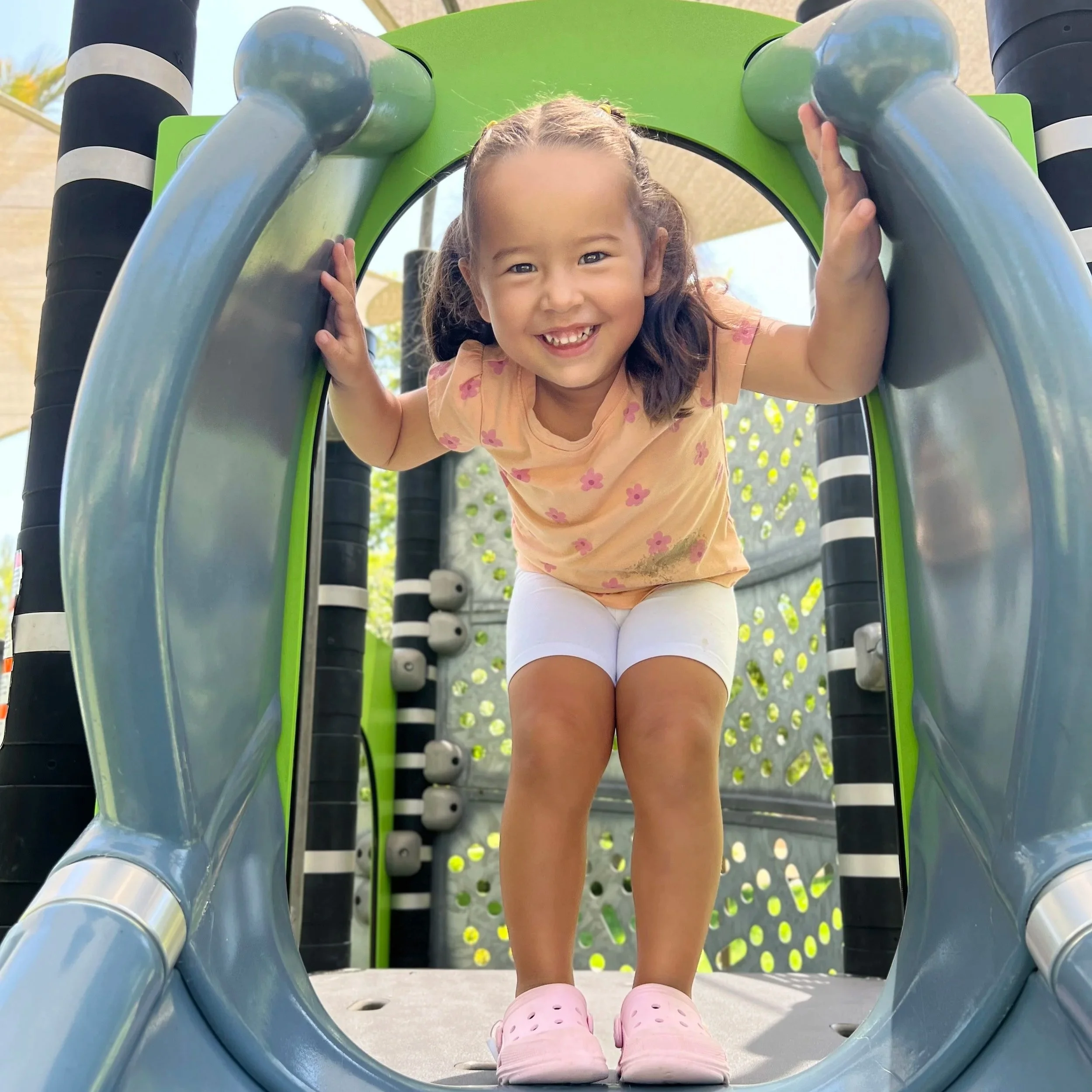 A young girl smiling and playing on a playground structure, crouching and holding onto the metal bars.