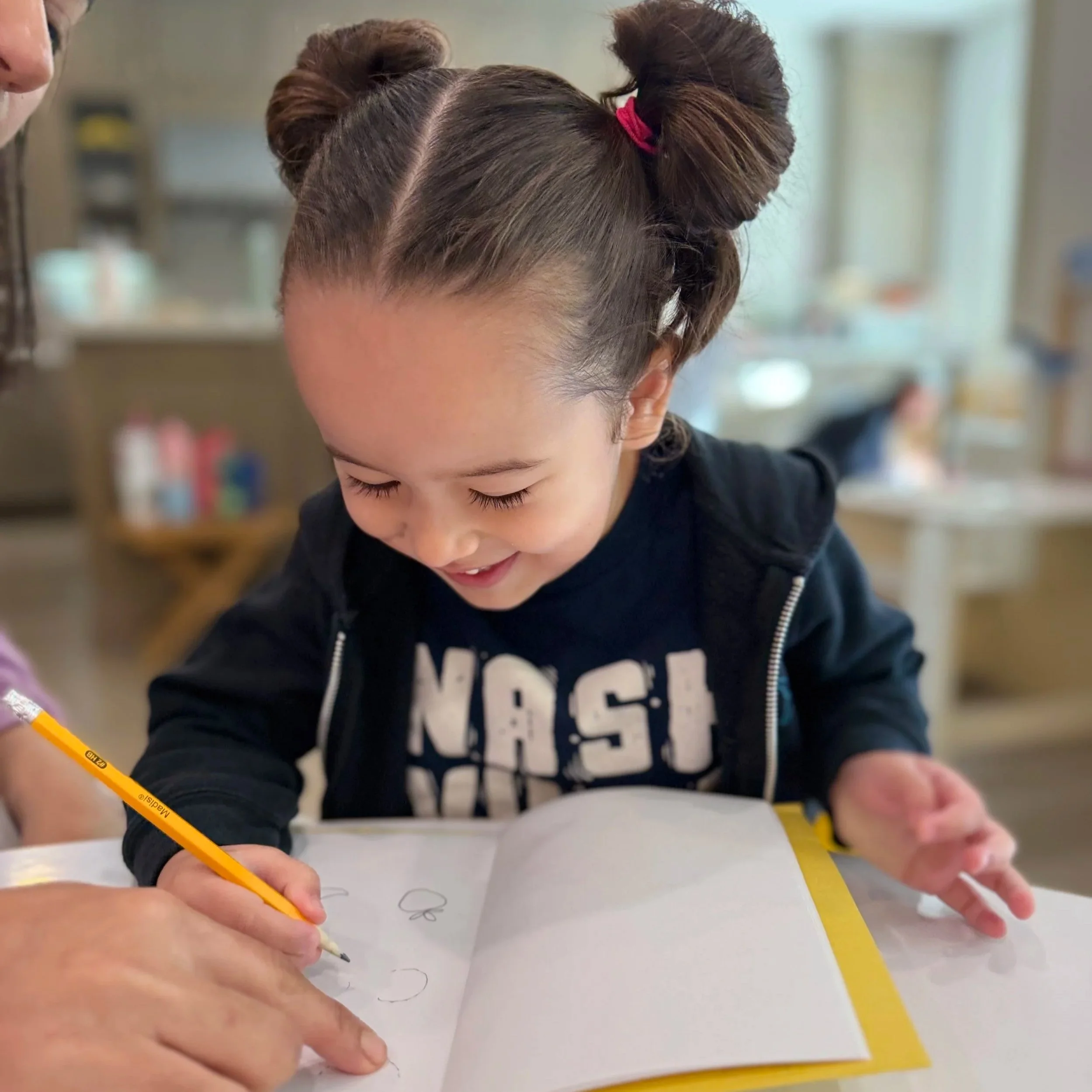 A young girl with brown hair styled in two buns, wearing a black jacket and a black shirt with 'NASA' printed on it, is smiling and drawing in a notebook with a yellow pencil. She appears to be in a bright room, possibly a classroom or art area, with blurred shelves and items in the background.