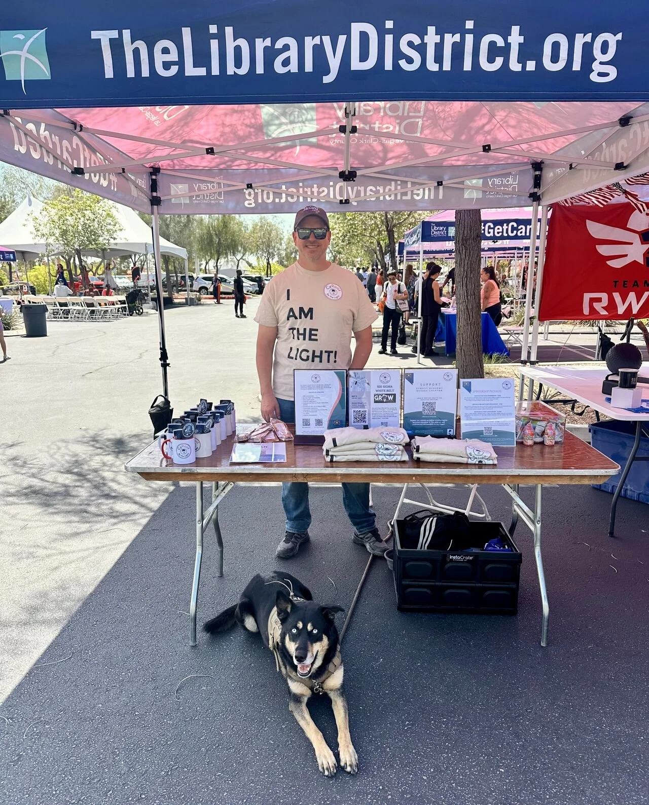 A man standing behind a table at an outdoor event, with a dog lying in front. The man is wearing sunglasses and a beige T-shirt that says 'I AM THE LIGHT!'. The table displays various items, and there is a blue canopy with the website 'TheLibraryDistrict.org' on it.