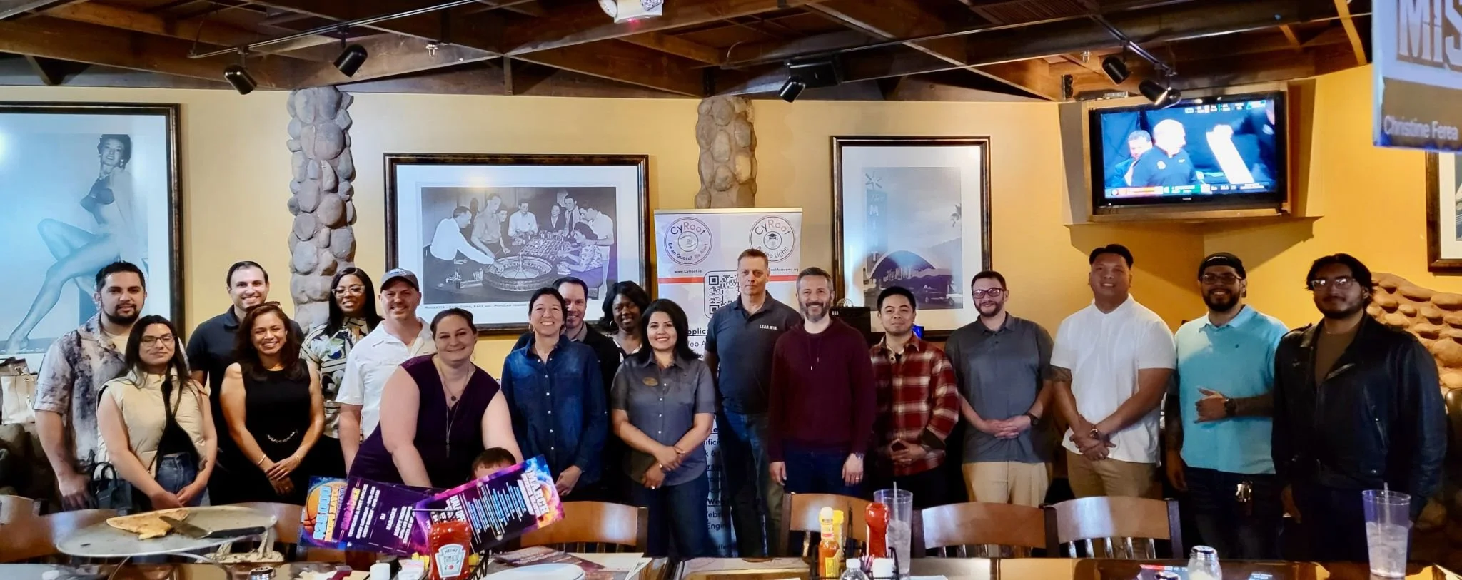 Group of people gathered for a photo inside a restaurant or bar with yellow walls, framed pictures, a stone pillar, and a TV. They are smiling and standing behind a table with drinks and condiments.