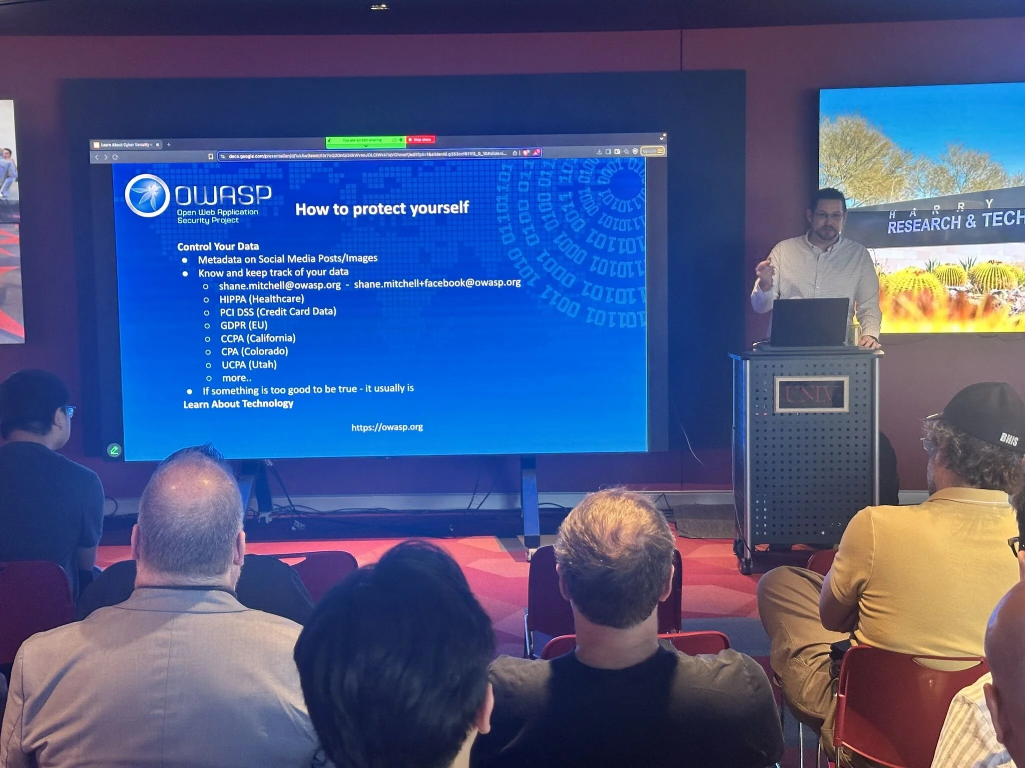 A man presenting a lecture on cybersecurity to an audience in a conference room. The presentation slide on the screen highlights how to protect oneself online, listing metadata control, tracking, and privacy guidelines.