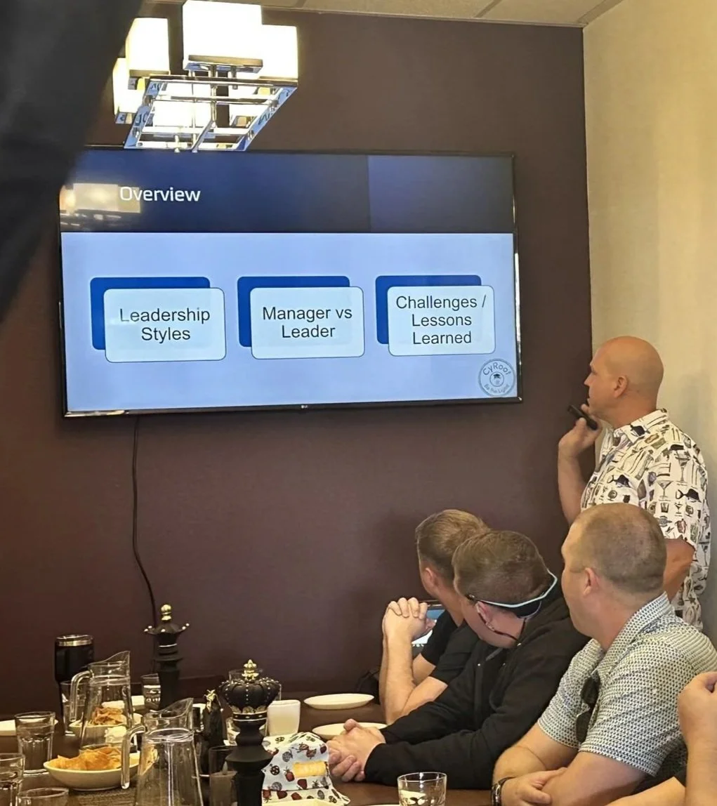 A man in a patterned shirt presents in front of a large screen displaying a slide titled 'Overview' with three points: Leadership Styles, Manager vs Leader, Challenges / Lessons Learned. Several people sit at a table with drinks and snacks, watching the presentation.
