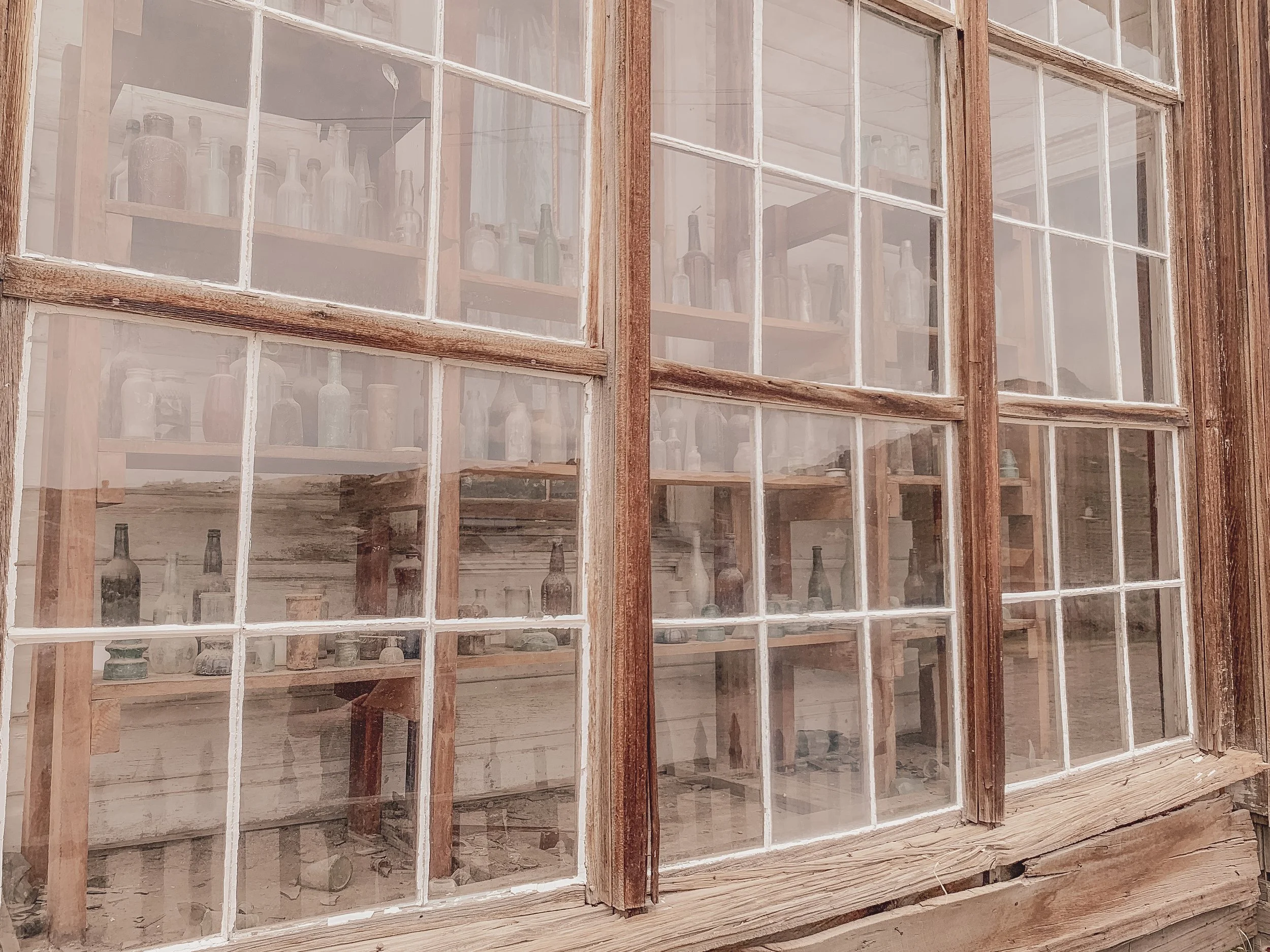 An old wooden window with multiple panes, showing a glass display case filled with bottles and containers inside a rustic building.