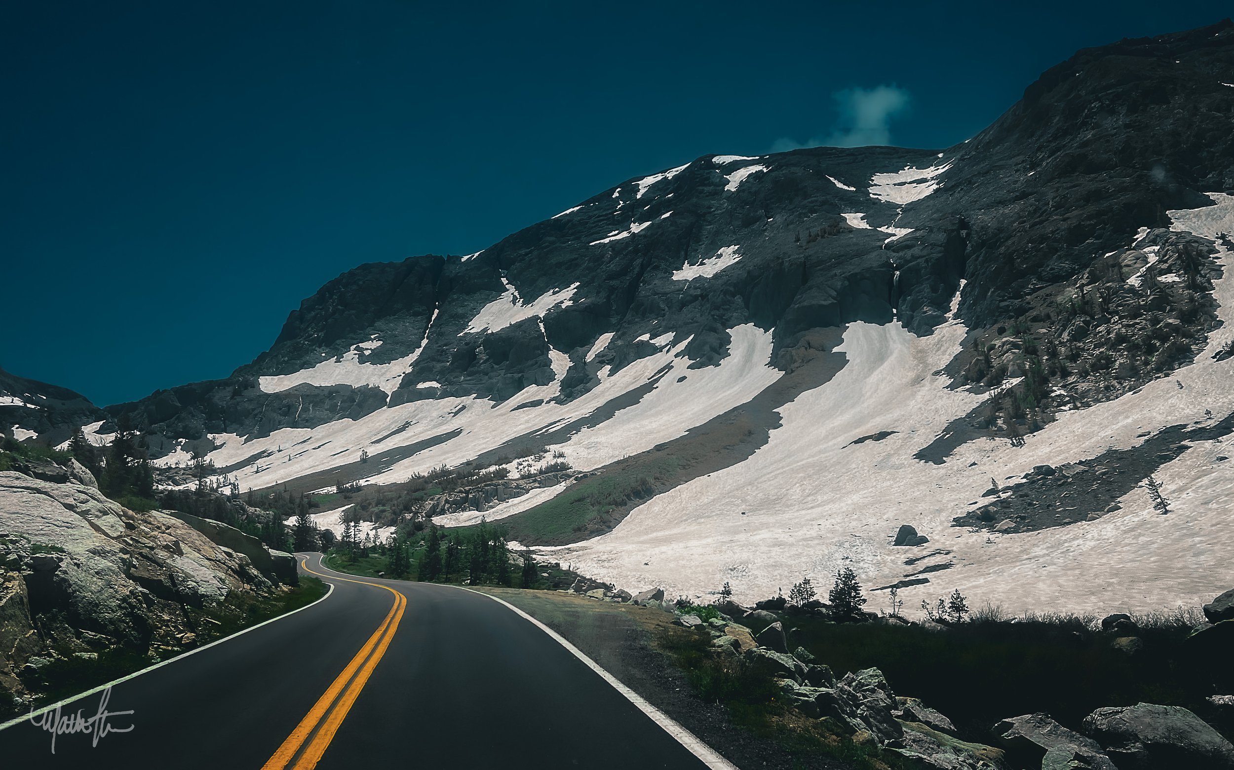 A winding mountain road with double yellow lines passing through a snowy mountainous landscape with rocks and evergreen trees.