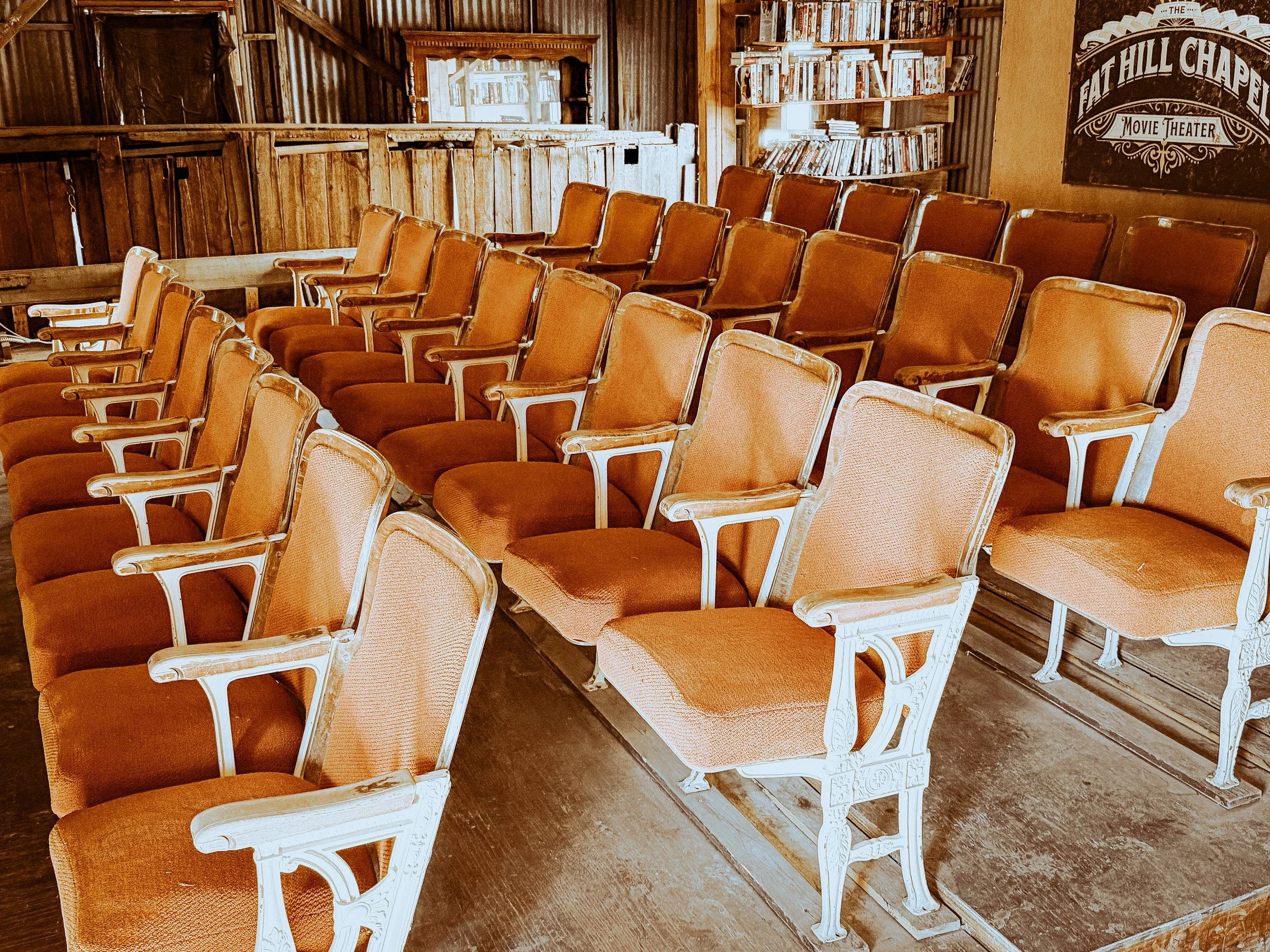 Row of empty vintage theater seats with wooden armrests and orange upholstery in a wooden interior, with bookshelves and a sign that reads 'The Flat Hill Chapel Movie Theater' on the wall.