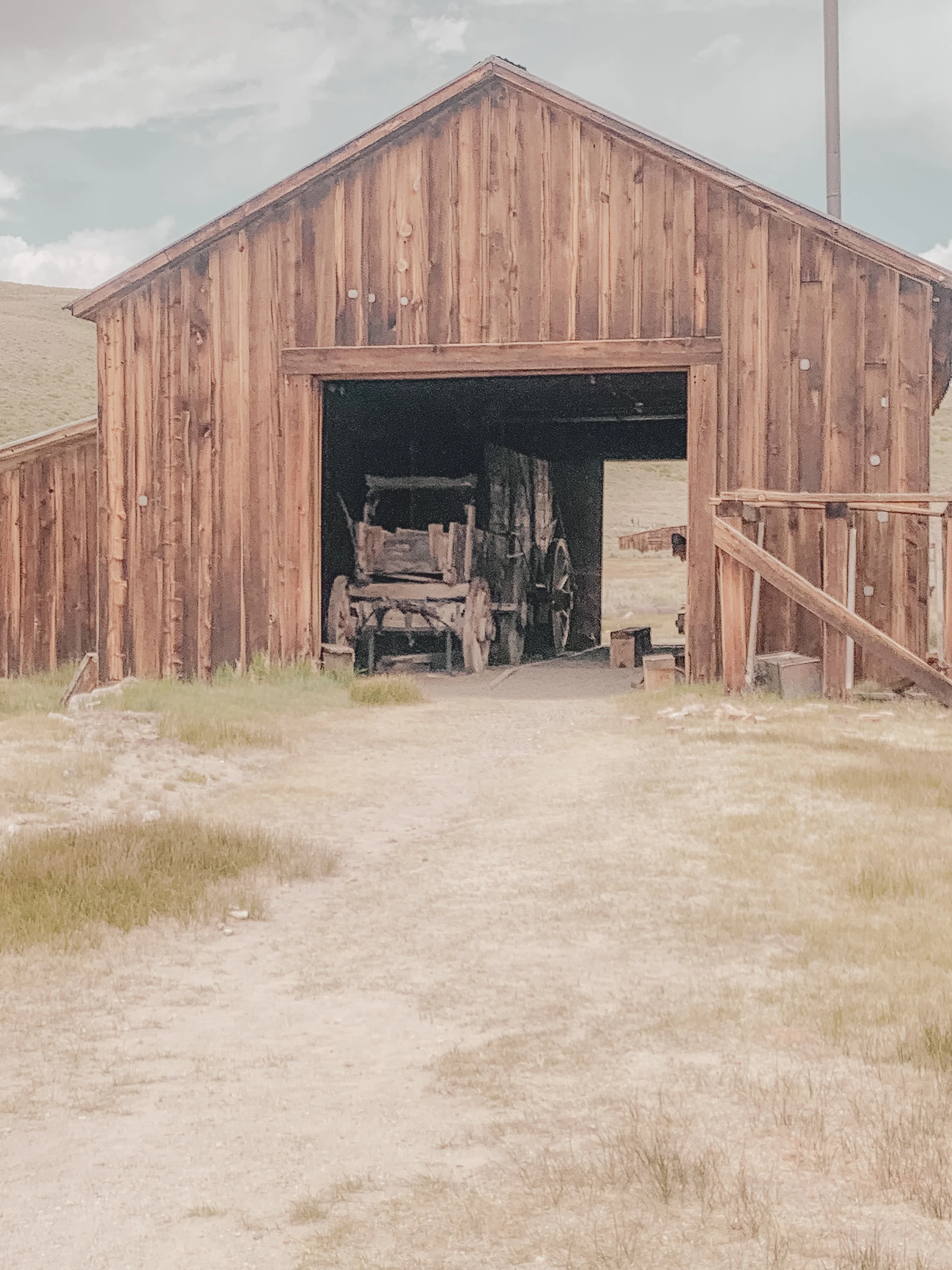 An old wooden barn with an open entrance showing an antique horse-drawn wagon inside, with a dirt and grassy path in front.