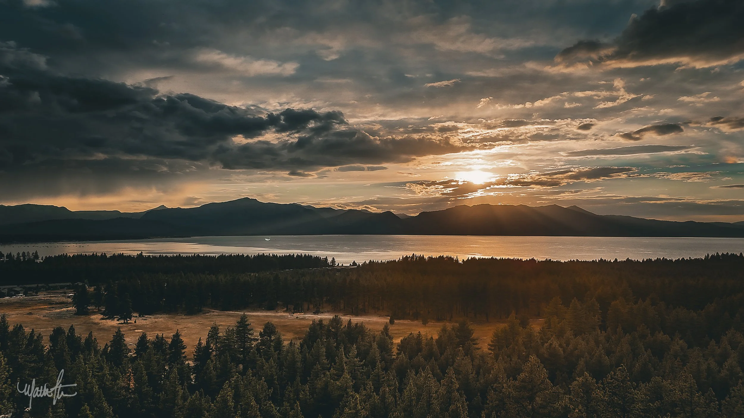 Sun setting over a mountain range with a lake in the foreground and a forest in the lower part of the image.