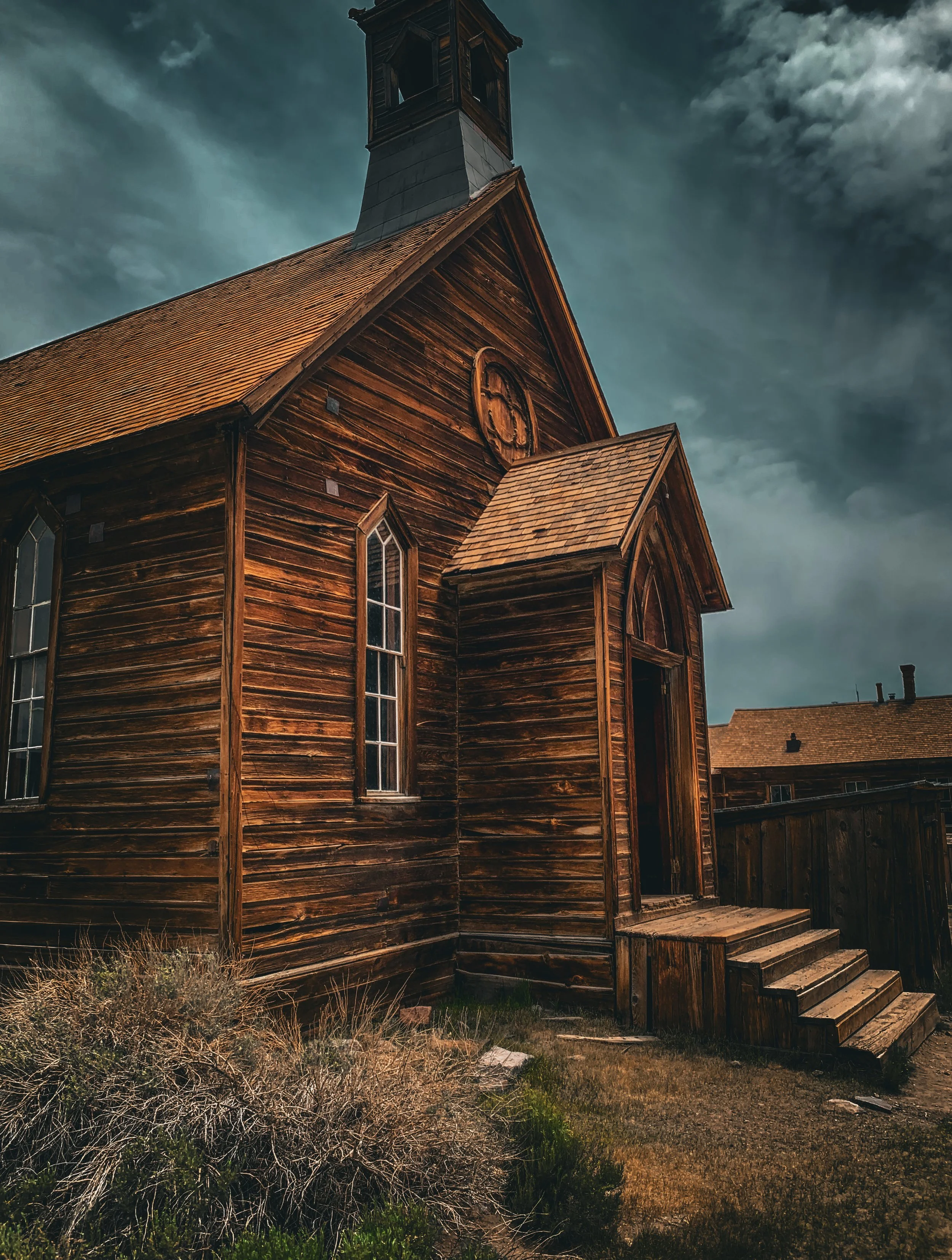 A wooden church with a steep roof, arched stained-glass windows, a small entrance with steps, and a bell tower against a cloudy sky.