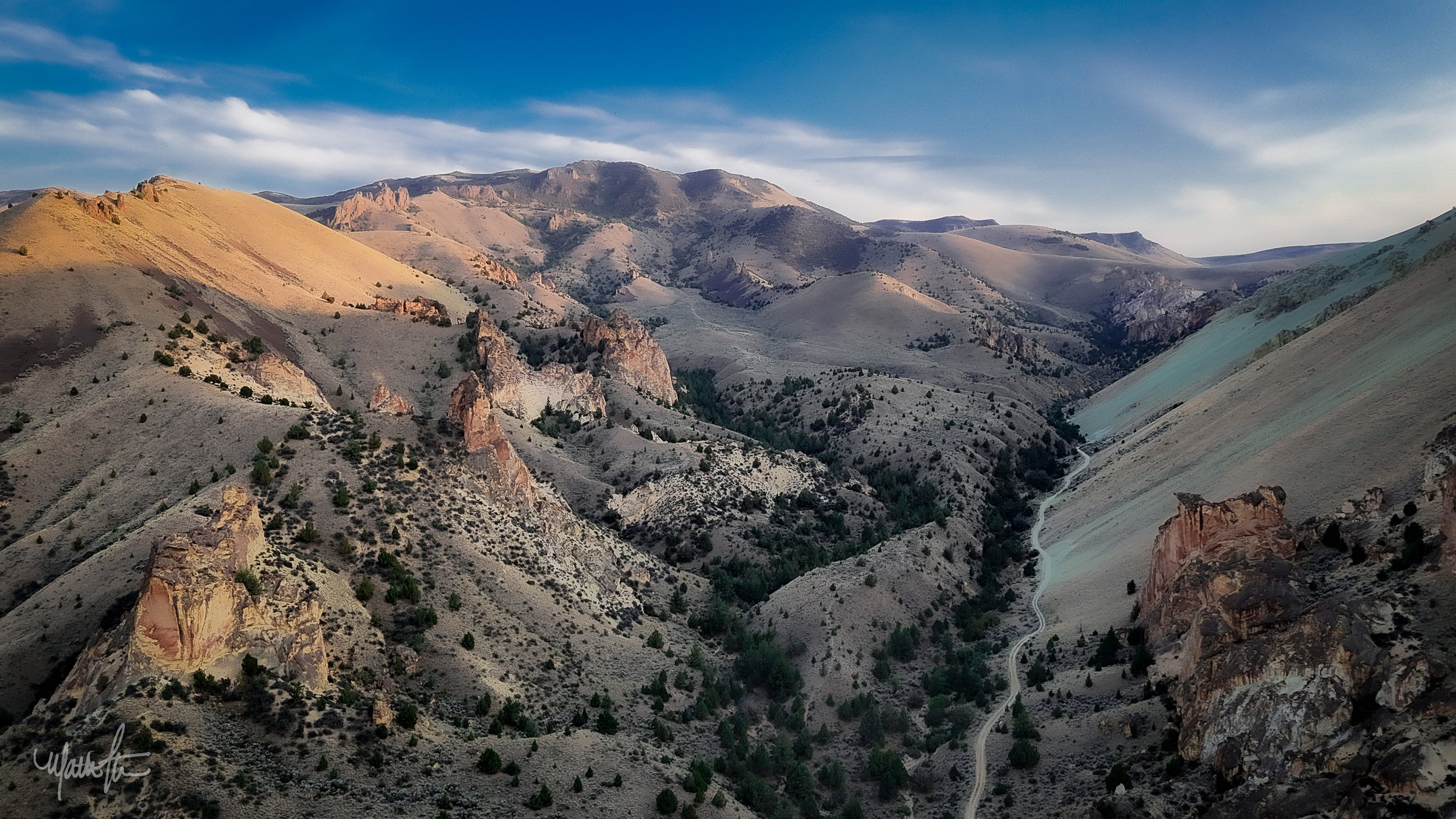A mountainous landscape with rugged terrain, sparse vegetation, and a winding road below. The sky is partly cloudy with blue sky visible.