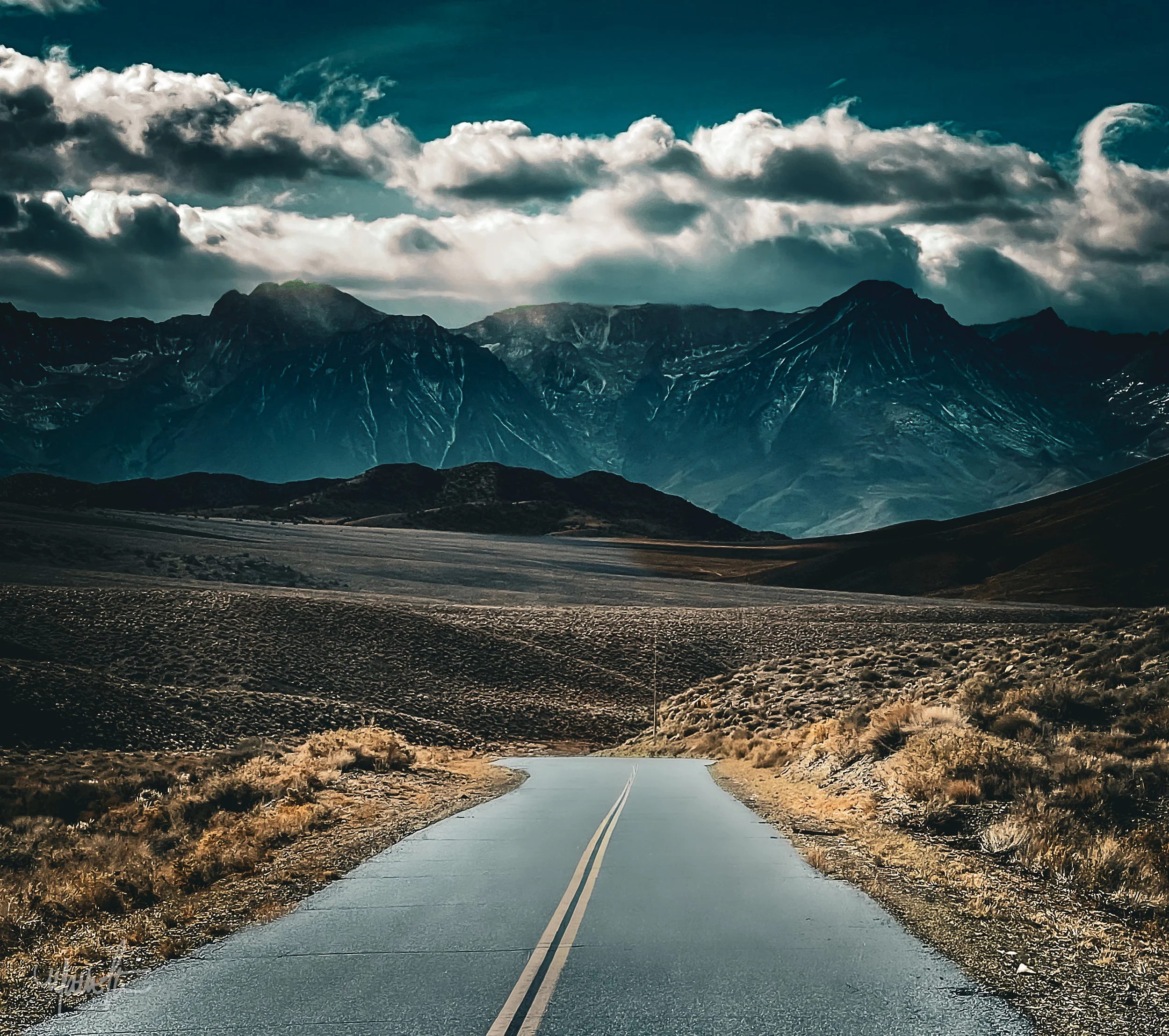 A person riding a motorcycle on a country road with mountains and dramatic clouds in the background.