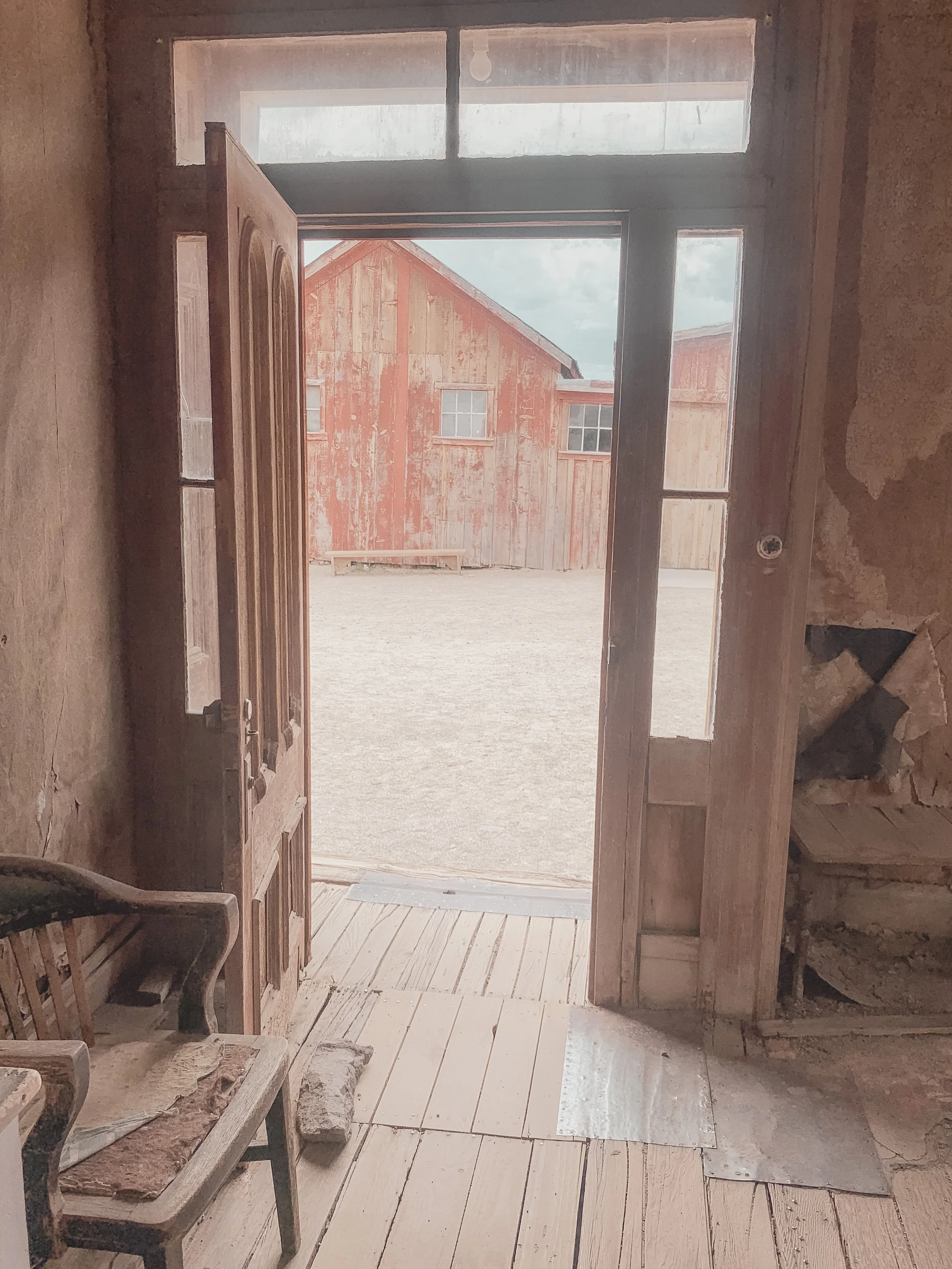 View through an open door showing a rustic red barn with weathered wood siding and small windows outside. Interior appears to be under renovation with unfinished walls and flooring.