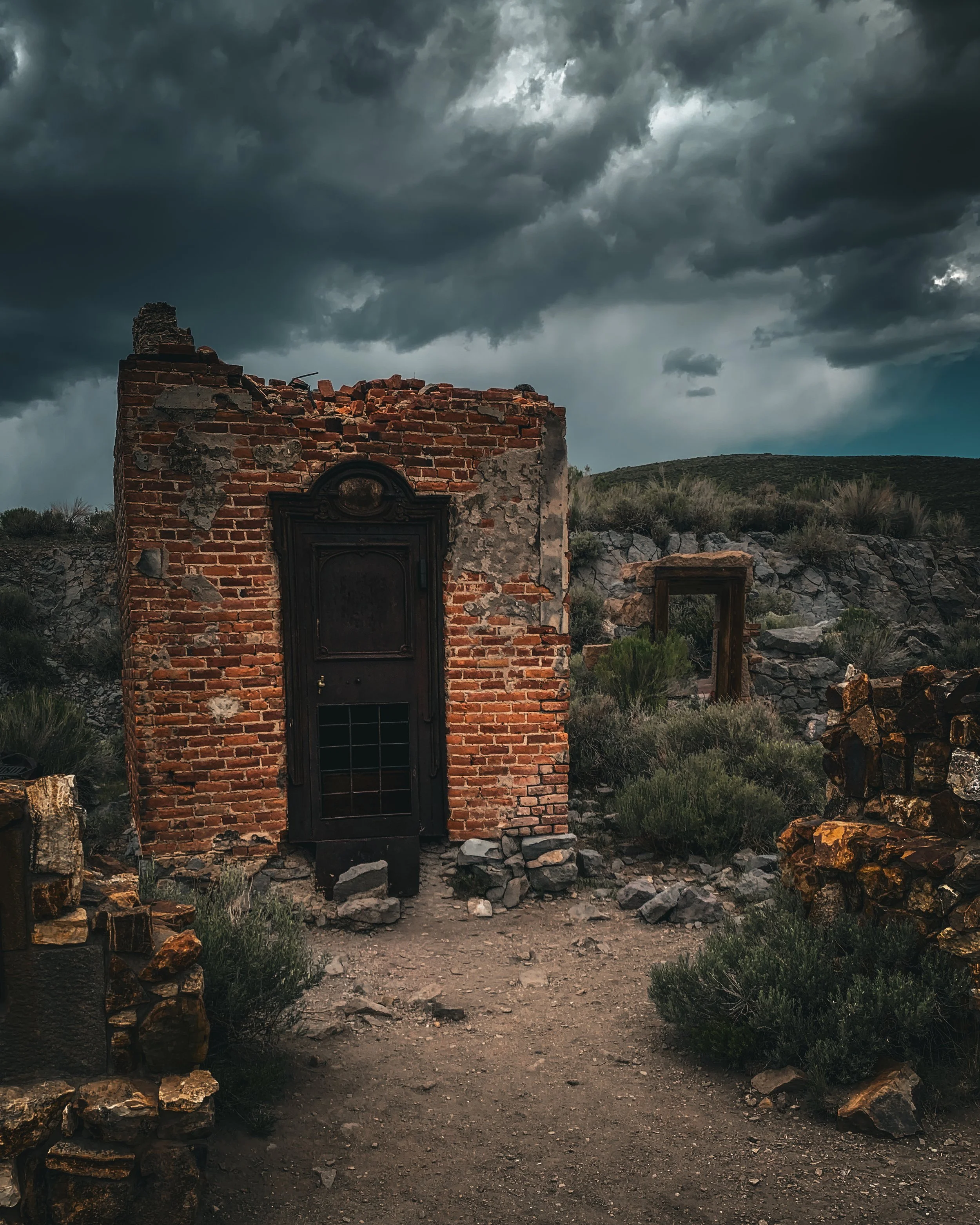 Ruined brick building with a black door amidst rocky desert terrain under dark stormy clouds.