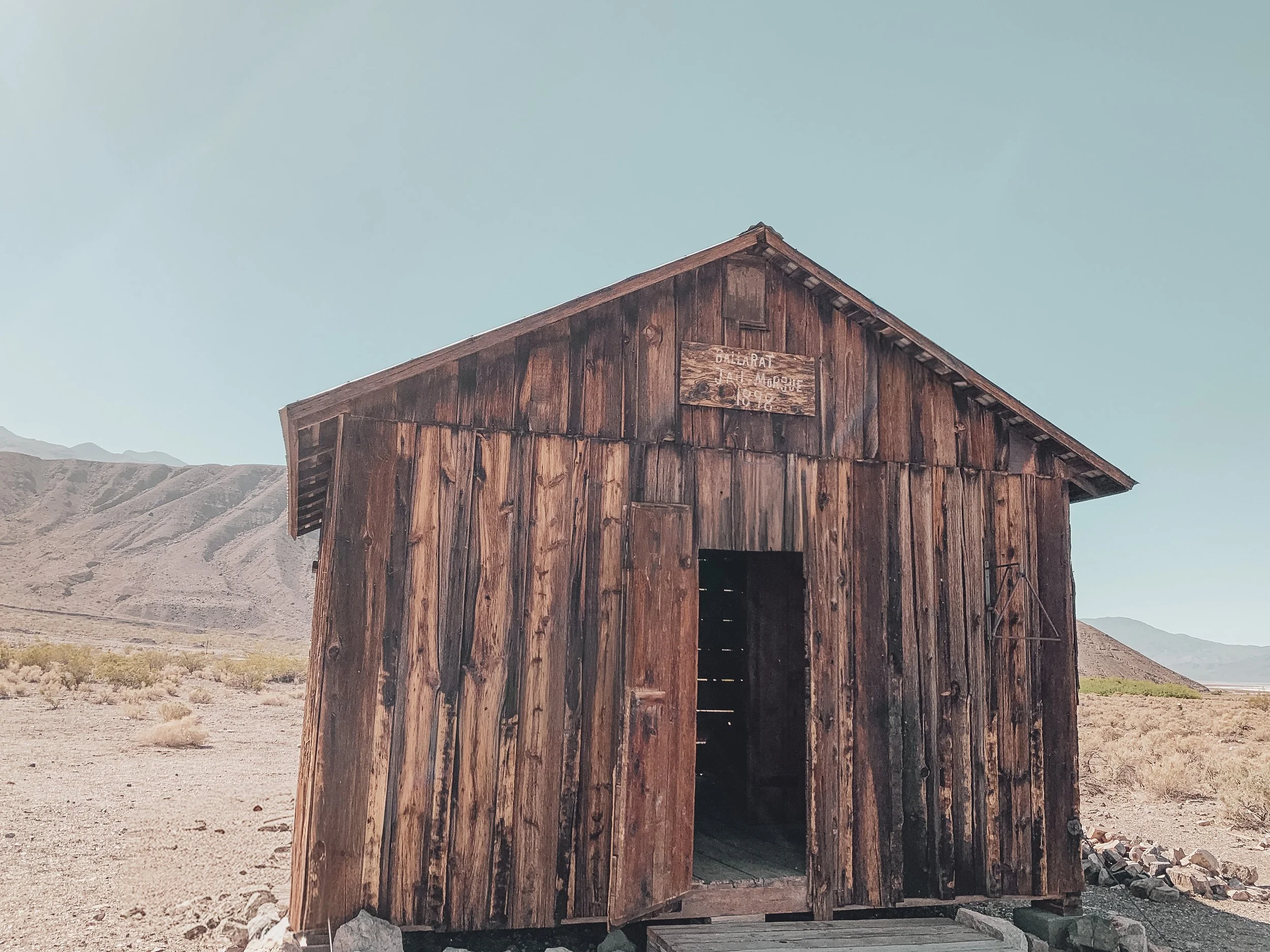 An old wooden barn in a desert landscape with mountains in the background and a clear blue sky.