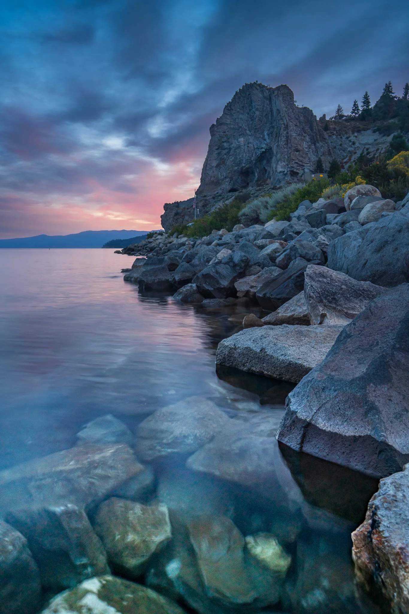 A rocky shoreline by a body of water with a large rock formation and a partly cloudy sky at sunset.