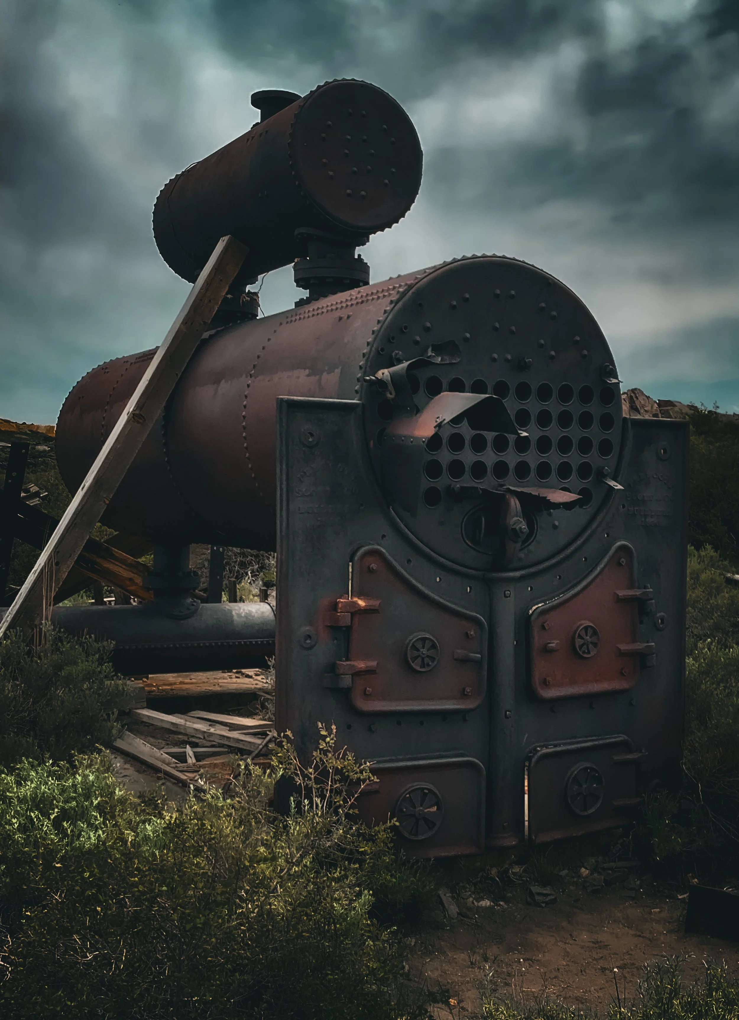 Old, abandoned industrial boiler or tank with rust and wear, set outdoors under a cloudy sky, surrounded by overgrown vegetation.