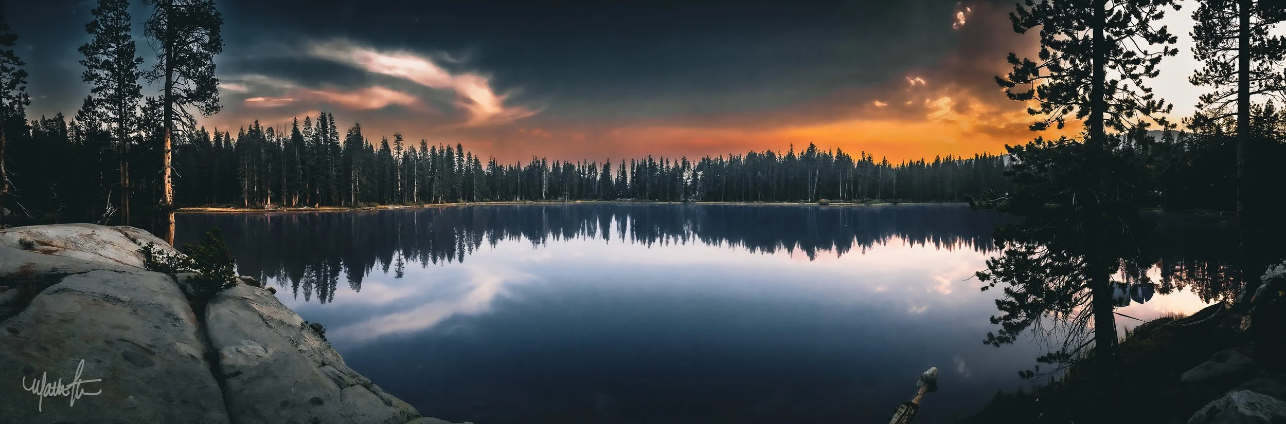 A tranquil lake surrounded by tall pine trees reflecting the sunset and cloudy sky.