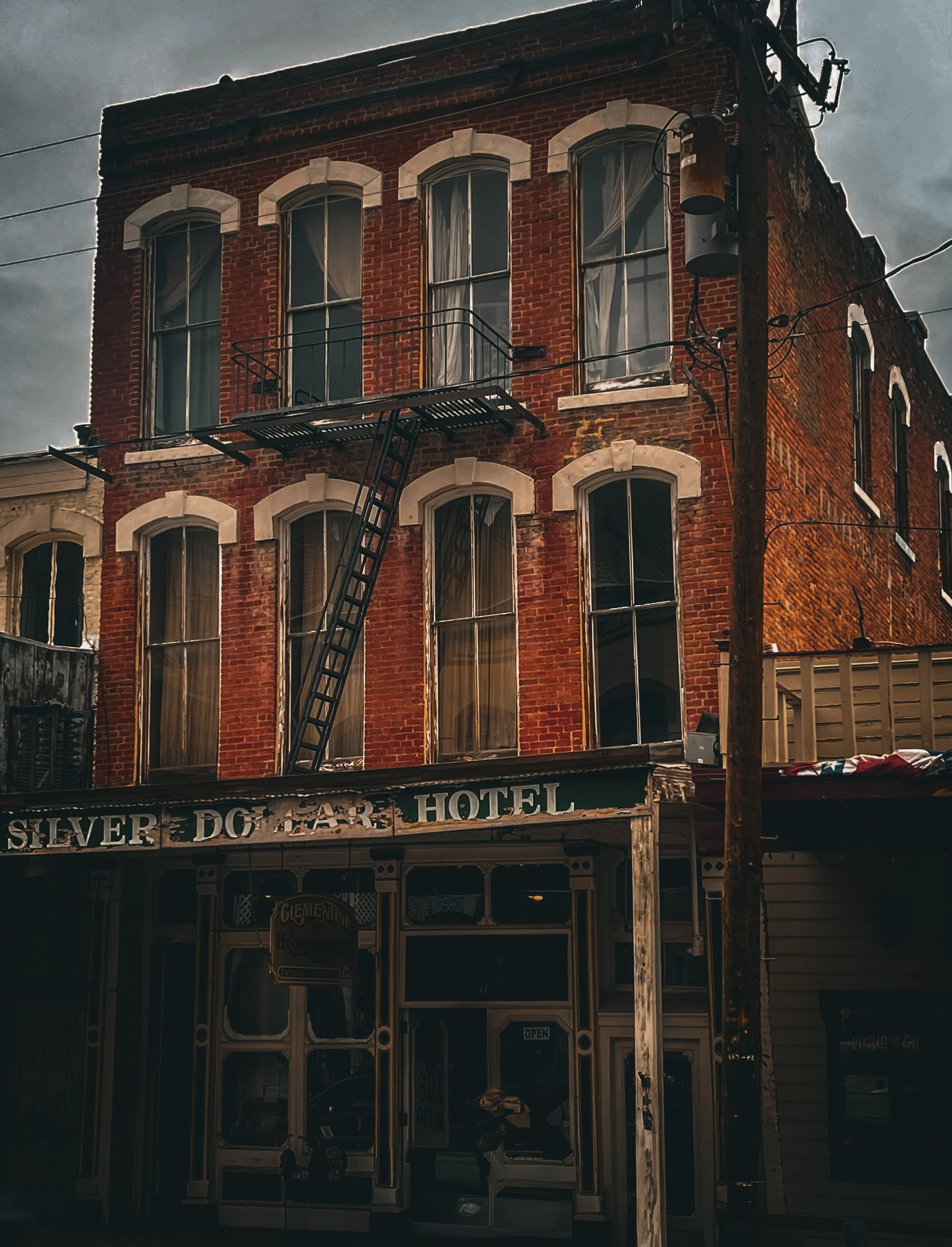 A three-story brick hotel building with tall windows, a fire escape ladder, and a weathered sign reading 'Silver Dog Hotel' in a vintage style. The entrance has large display windows and an open sign. The sky is cloudy.