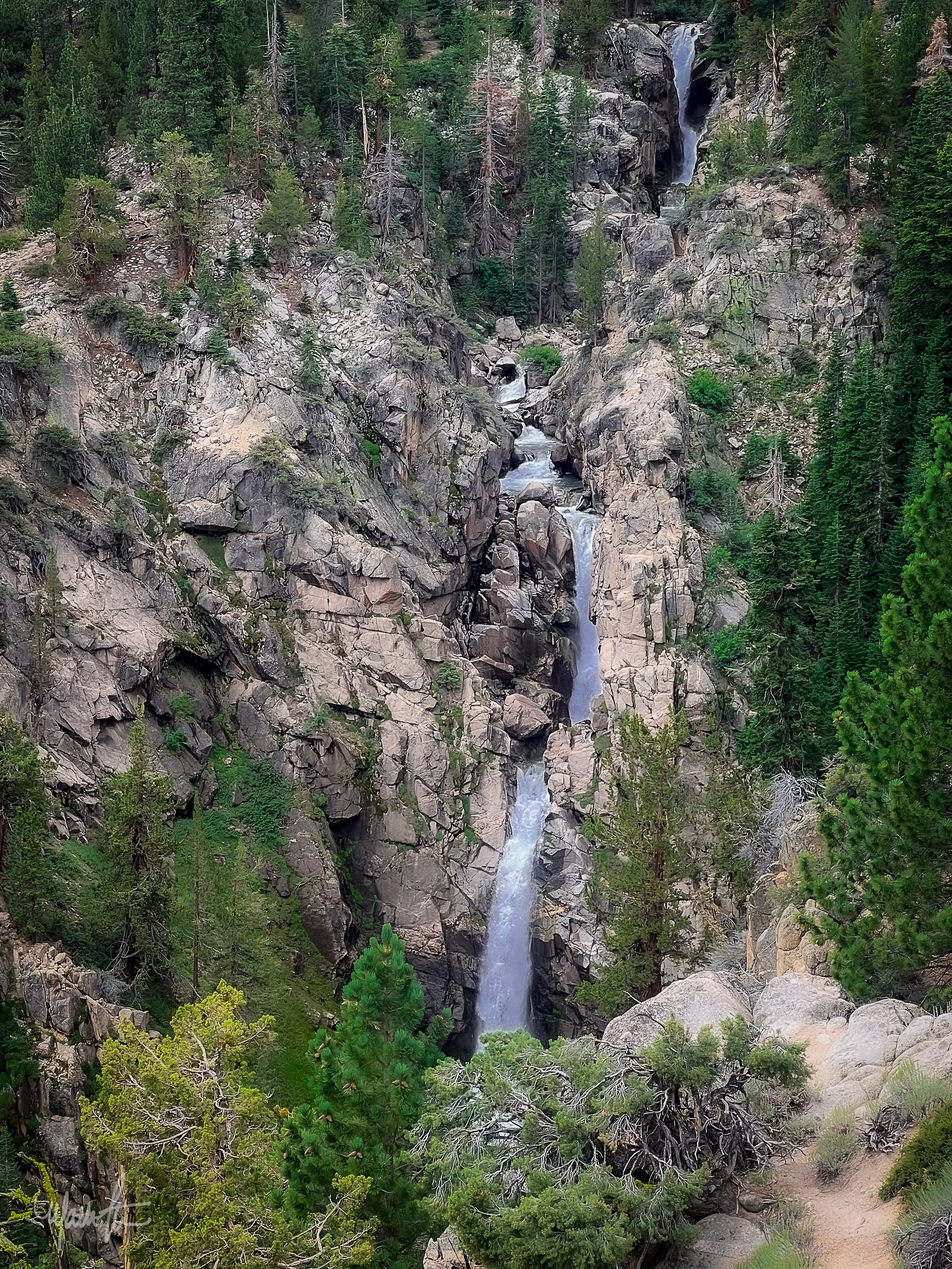 A tall, narrow waterfall cascading down a rocky cliff surrounded by lush green trees.