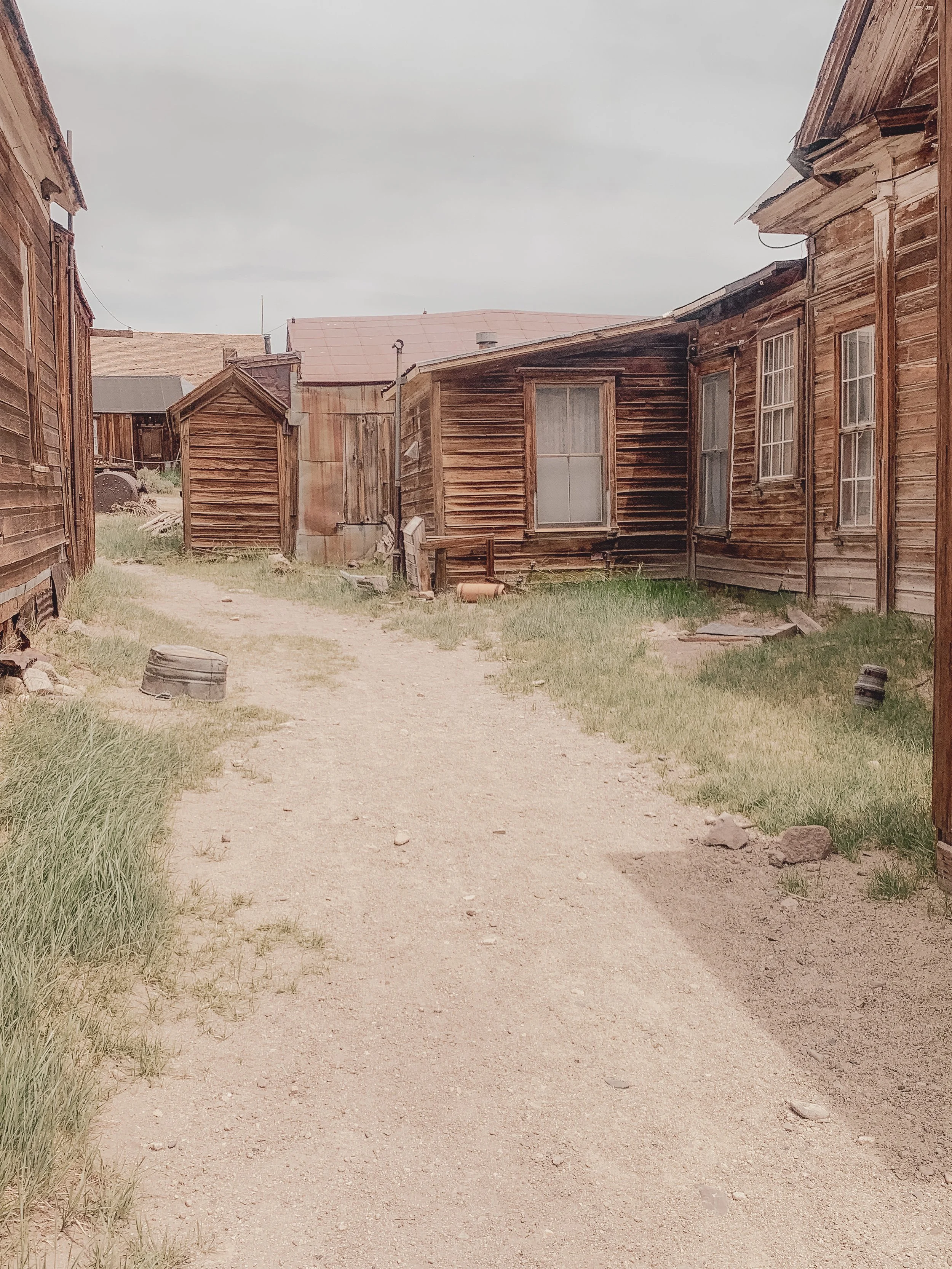 Old wooden buildings along a dirt path with grass on sides and an overcast sky.