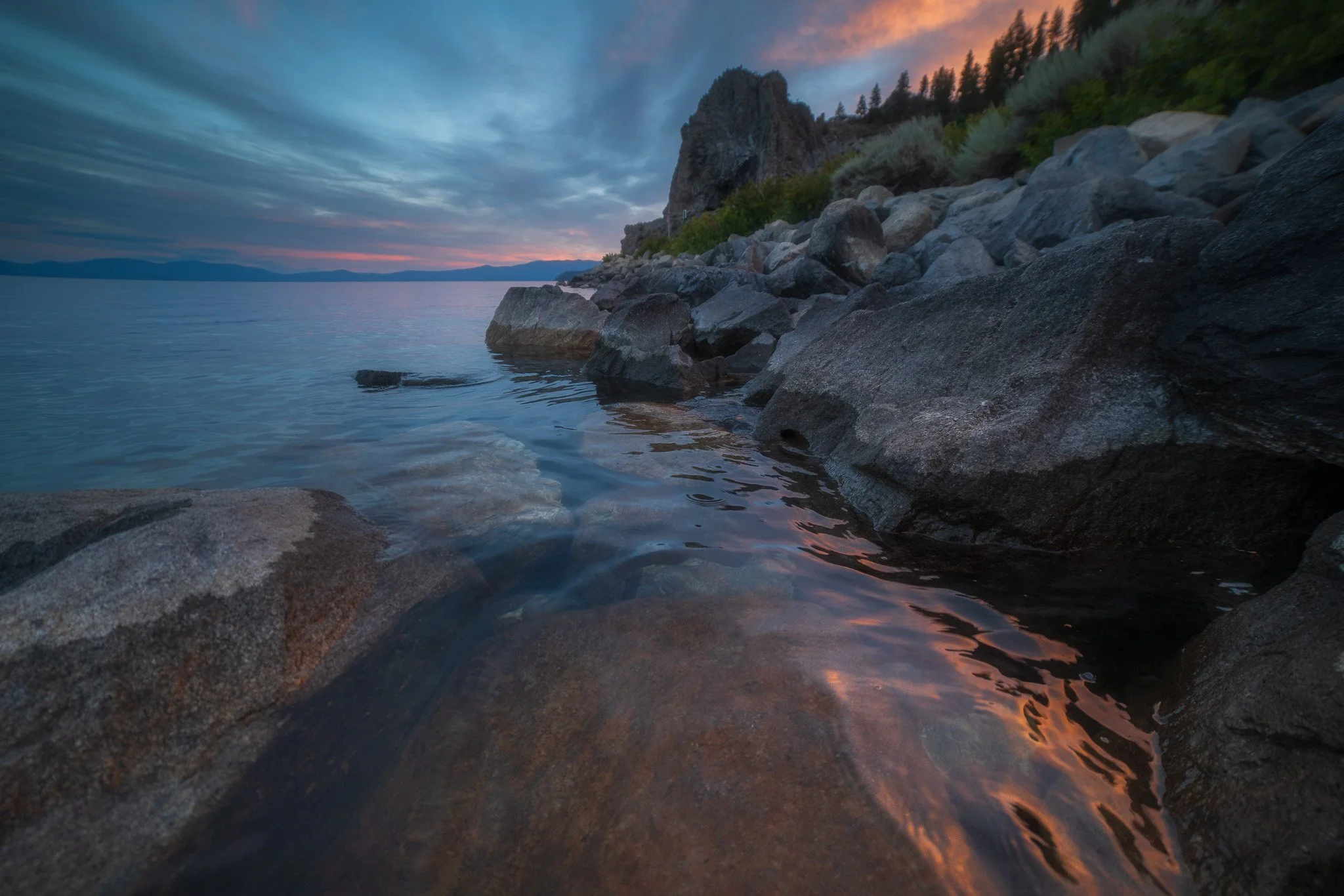 A rocky shoreline at sunset with calm water, a cloudy sky with hints of pink and orange, and a distant mountain range.