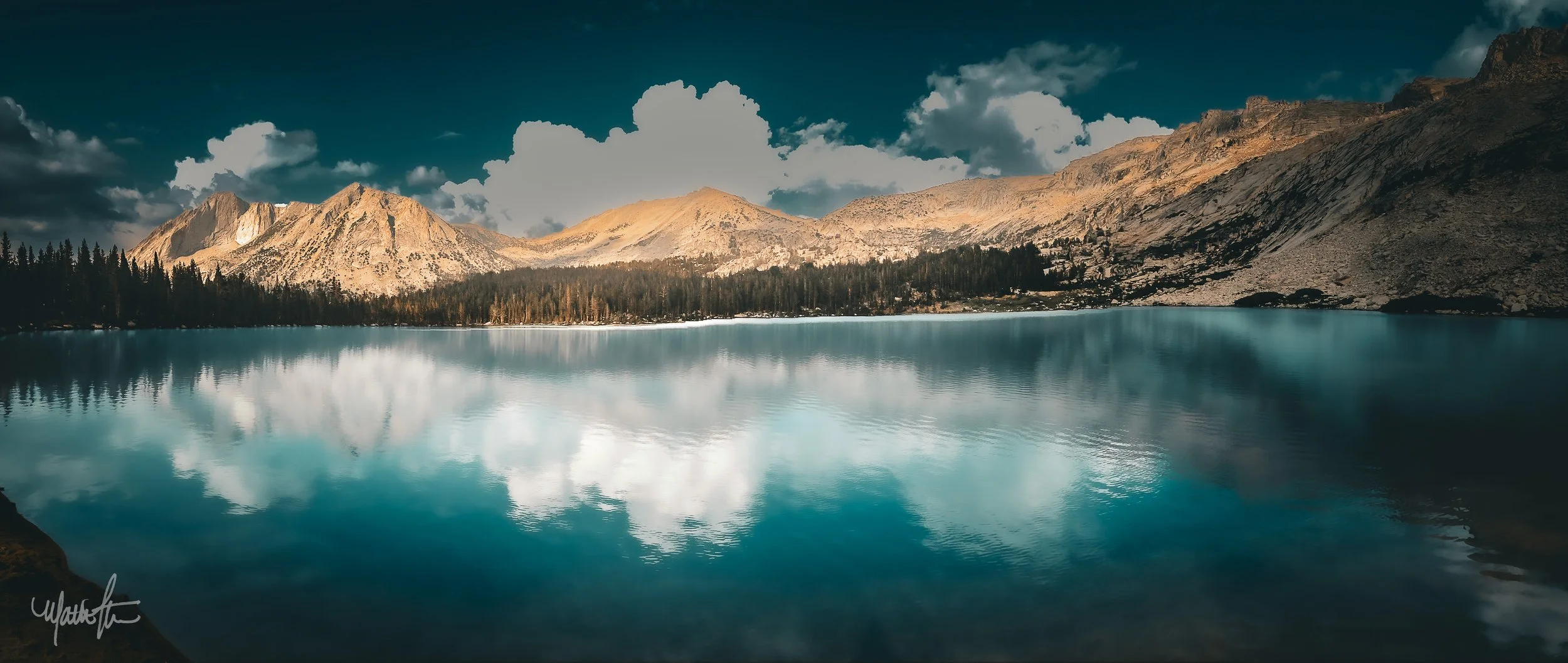 A serene mountain lake surrounded by pine trees, with rugged mountains in the background under a partly cloudy sky.