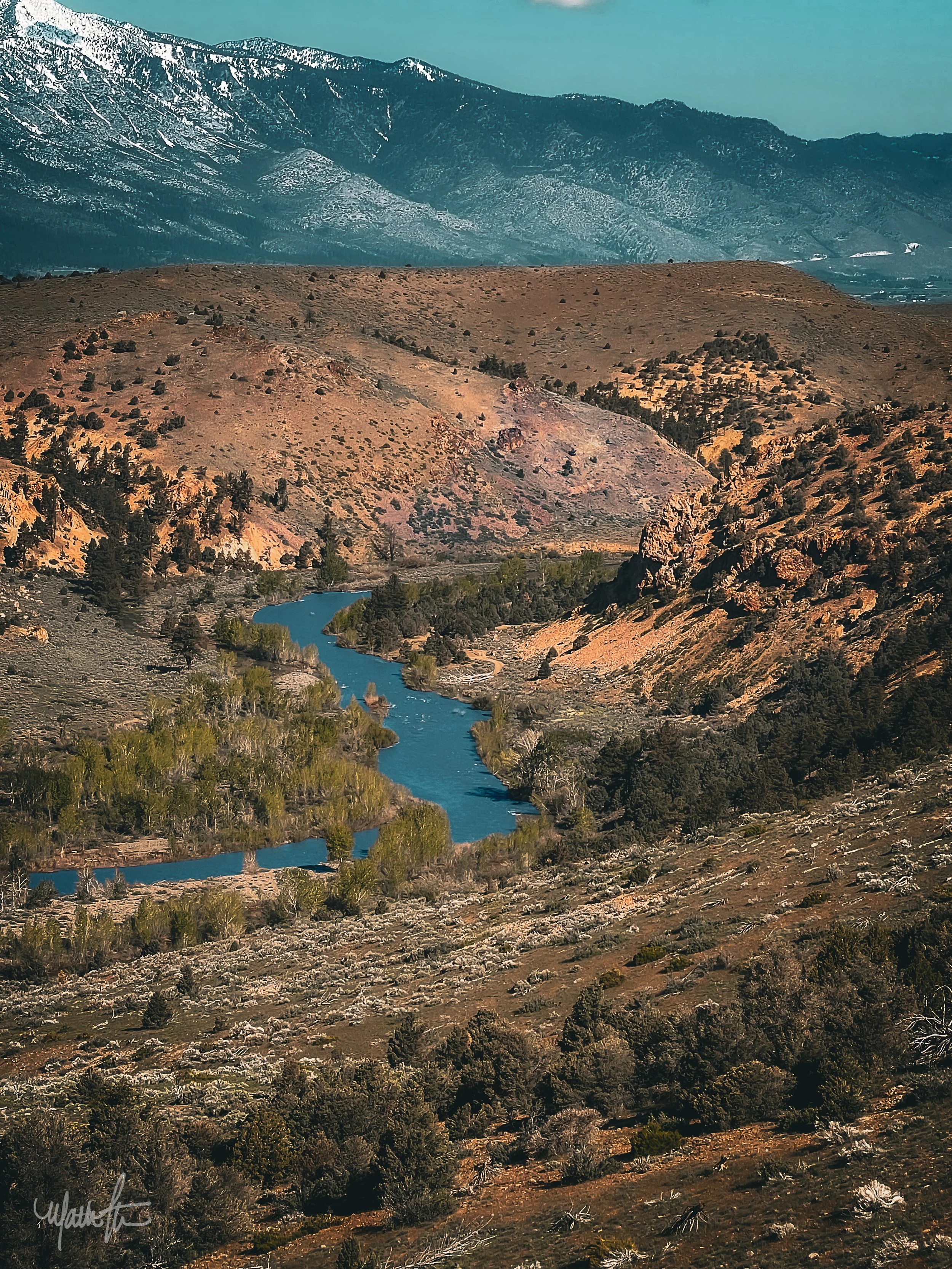 A winding river flowing through a mountainous landscape with sparse trees and rugged terrain, snow-capped mountains in the background.