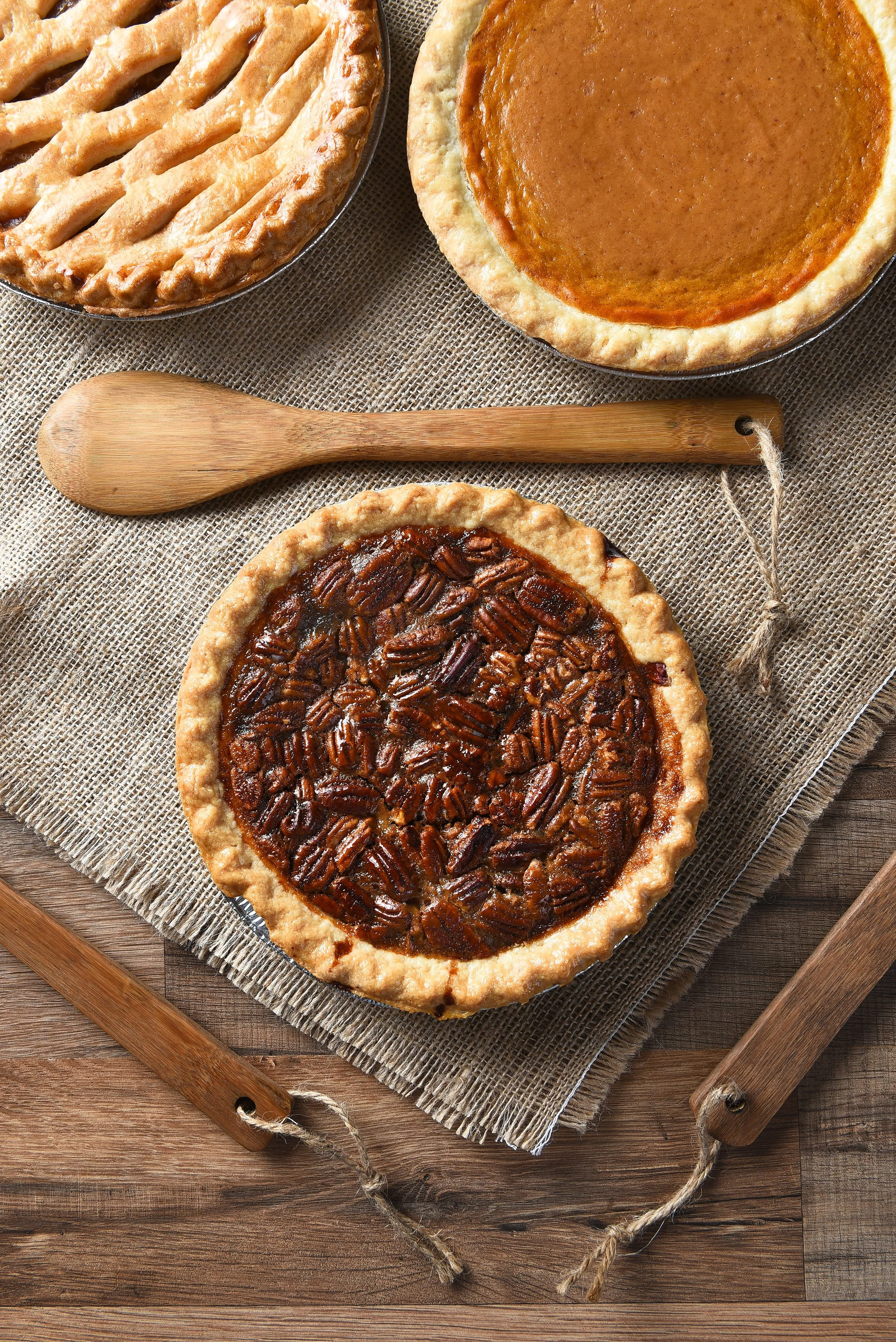 Three different types of pies on a rustic wooden surface, including pecan, pumpkin, and sweet potato, with a wooden pie server nearby.
